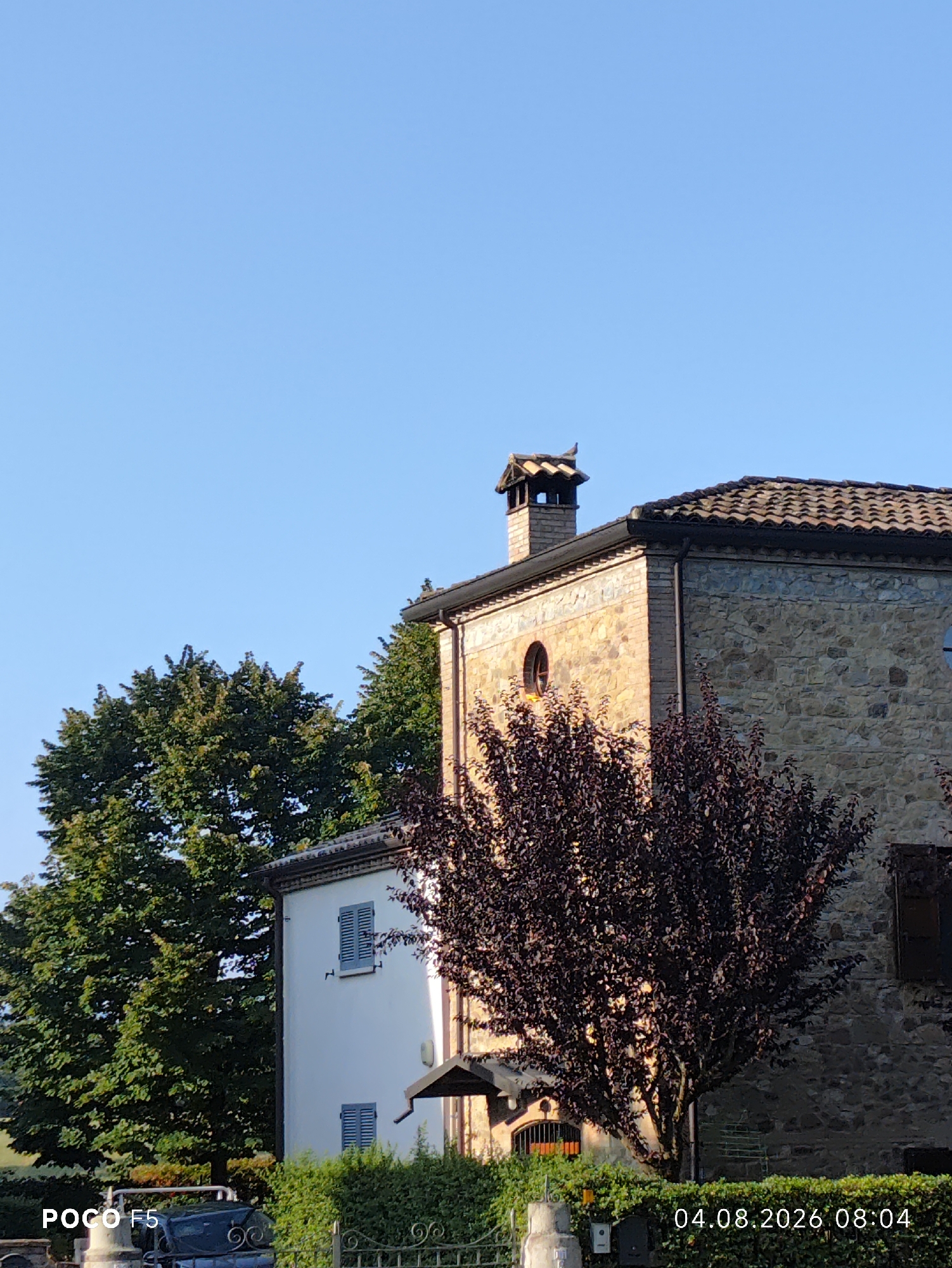 A rustic stone and brick building, partially obscured by trees, stands under a clear blue sky. The building features a tiled roof, a chimney, and a circular window. To the left of the main building is a white structure with blue shutters. A dark-leaved tree is in the foreground, and lush green trees are to the left. The scene is set in an outdoor environment, possibly a rural or suburban area. The lower left corner displays "POCO F5," and the lower right corner shows "04.08.2026 08:04," suggesting the image was captured on August 4, 2026, at 8:04 AM.