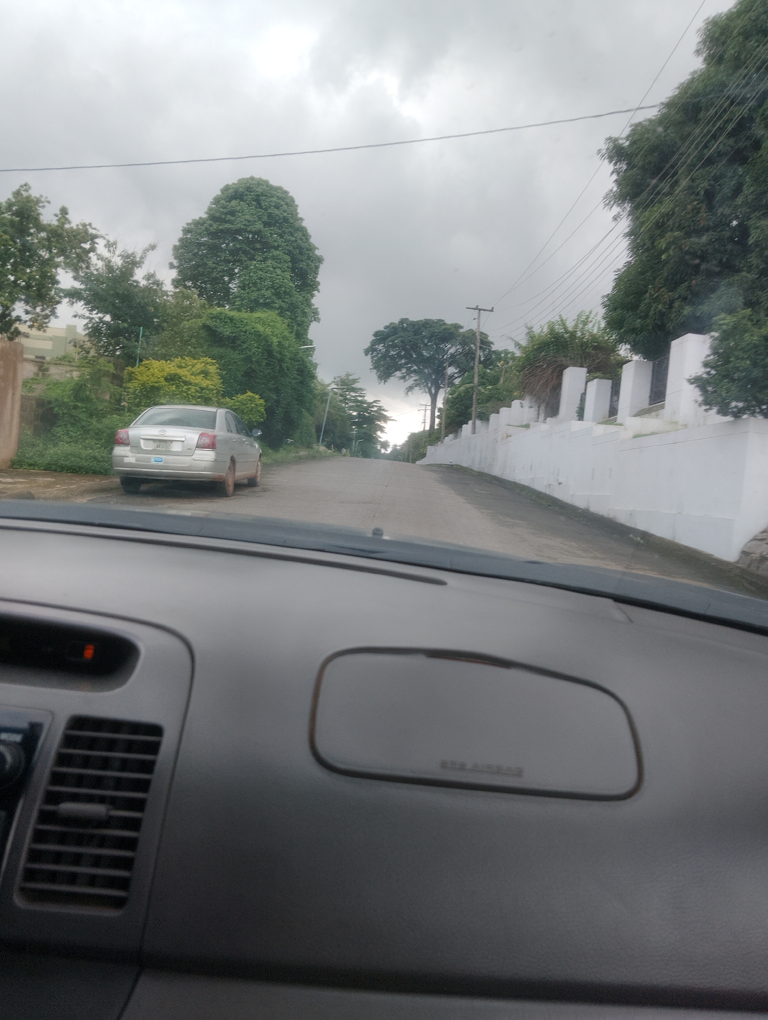 A silver sedan is parked on the right side of a paved road that curves upwards and to the right. The car faces away from the viewer and towards the right. A white wall with regularly spaced pillars runs along the right side of the road. Green foliage and trees are visible on both sides of the road, with a larger, mature tree on the left. The sky is overcast and grey. The scene is viewed from inside a vehicle, with the dashboard and part of the windshield visible in the foreground. The image appears to be taken in Iju, Nigeria.