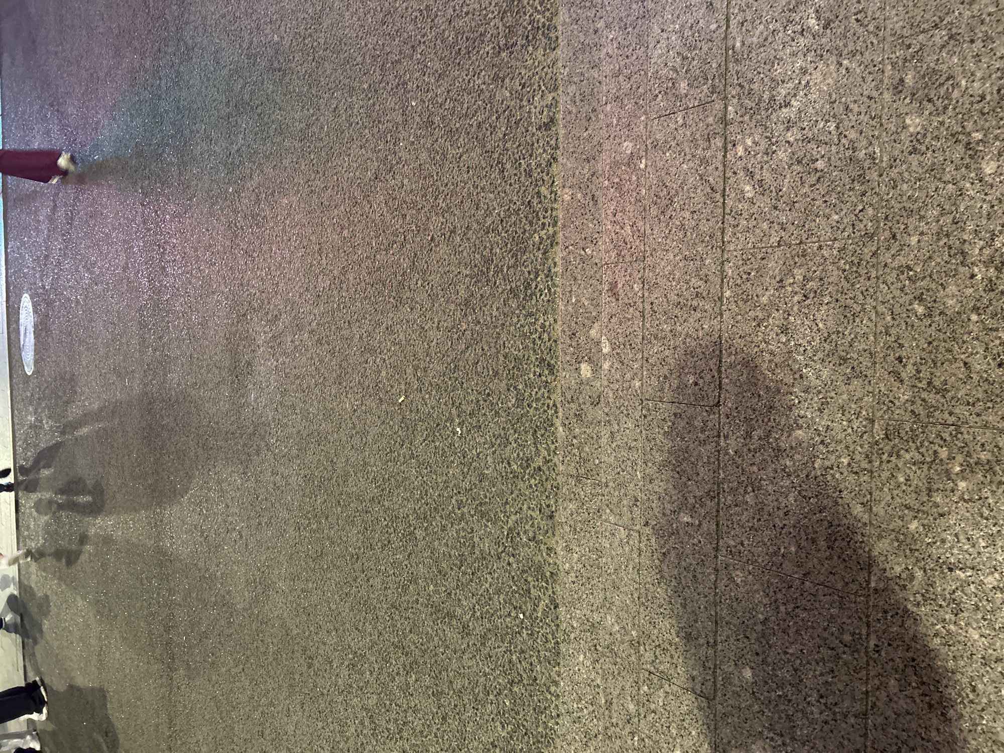 The image shows a street scene in an urban environment. The ground is paved with two different materials: one is a rough, dark asphalt, and the other is a smoother, grey stone tile surface. The asphalt section appears to be wet or reflective, as it picks up light with a pinkish hue. There are vague shadows and silhouettes of people's legs and feet on the left side of the image, indicating foot traffic. A small section of what may be a sign or marker is visible on the ground near the people. There is a dark shadow cast on the stone tiled part.