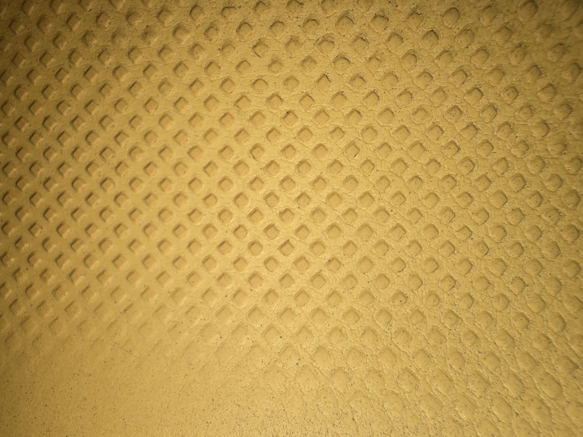 The image shows a close-up, abstract view of a textured yellow surface. The texture consists of a repeating pattern of small, raised squares arranged in a grid. The lighting creates shadows that emphasize the three-dimensionality of the pattern. The color is a consistent, muted yellow, with some slight variations in tone due to the texture and lighting. There are no discernible people, actions, or interactions. The image is solely focused on the surface itself. There is no contextual information available to place this image in the city of Ulan-Ude, Russia.