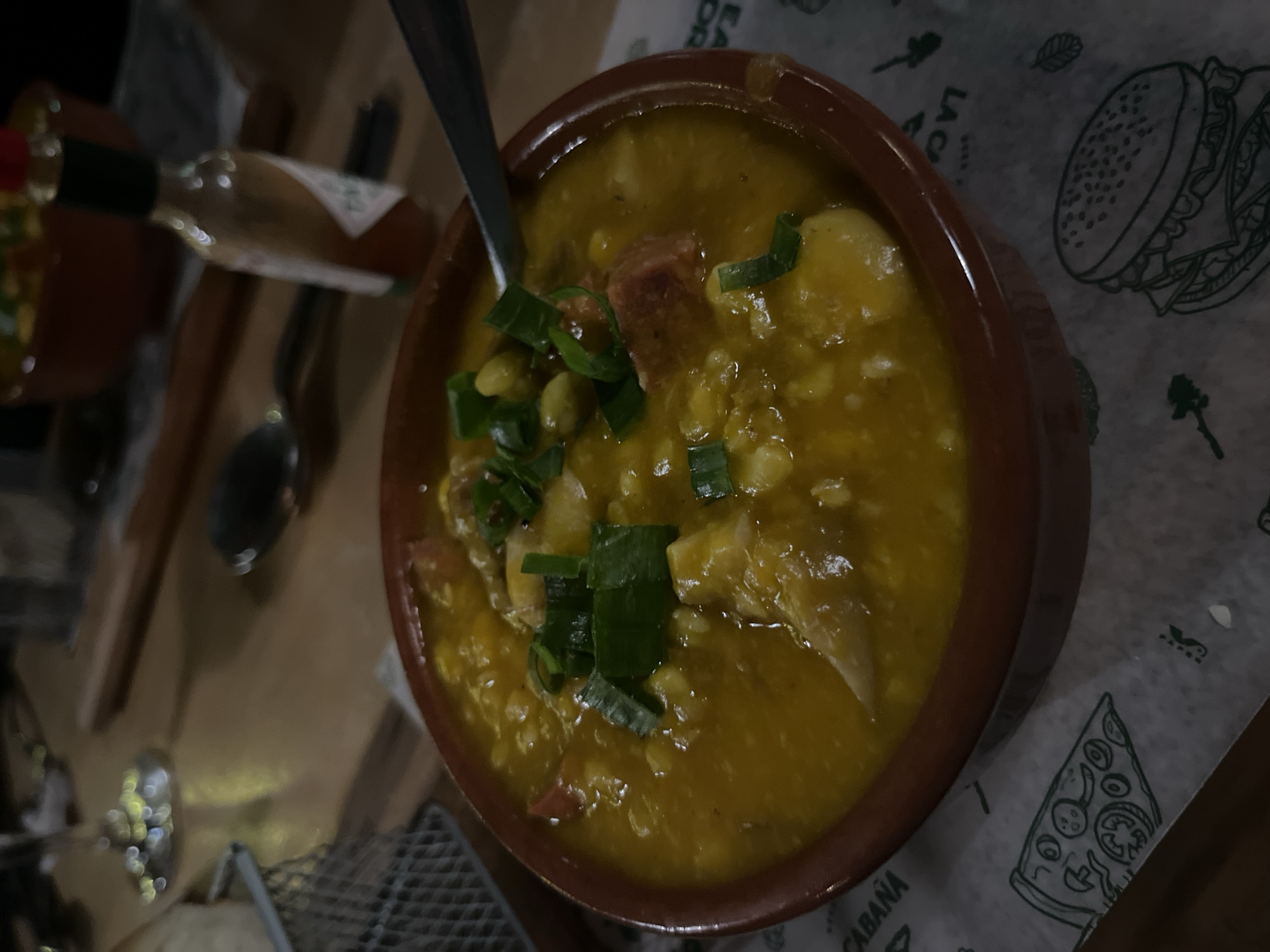 The image depicts a bowl of Locro, a thick stew popular in the Andean region, garnished with chopped green onions and containing visible pieces of meat, beans, and corn. A spoon rests in the bowl. The bowl sits on a branded paper liner with illustrations of a burger and pizza slice. A bottle of Tabasco sauce and eating utensils are nearby. The setting is most likely a restaurant. Due to the presence of Locro on the menu, the location is probably somewhere in Argentina, possibly in the city of Ushuaia, Argentina.
