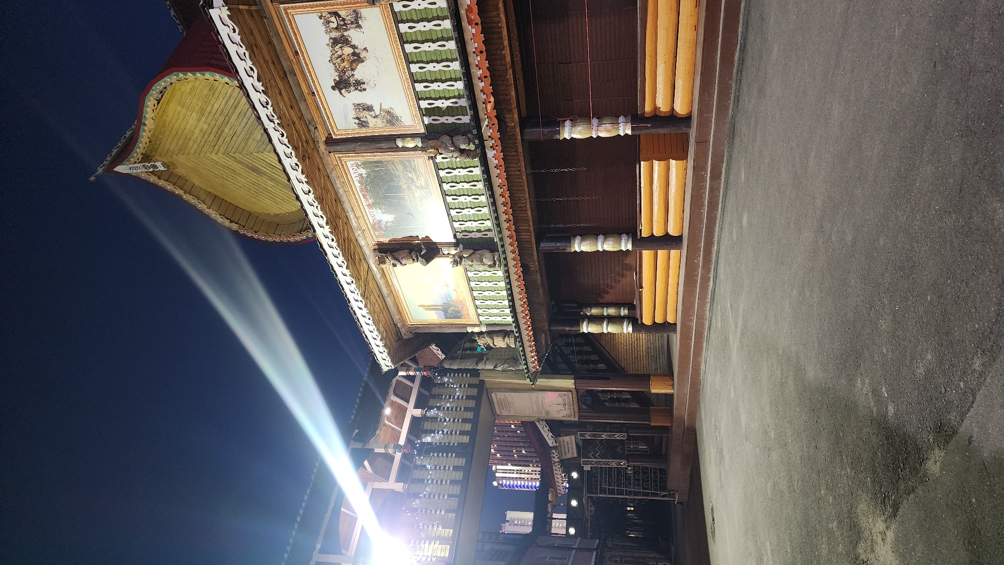 The image depicts a nighttime exterior view of a traditional wooden building complex, likely a historical or cultural attraction, in Moscow, Russia. The architecture features ornate carvings, decorative trim, and large framed paintings displayed on the facade. The building has multiple levels, including balconies and walkways, with a prominent, curved roofline adorned with intricate patterns. Logs are stacked in horizontal layers to form a lower portion of the structure. In the foreground, a paved courtyard area is visible, with some areas appearing wet or reflective, suggesting recent precipitation. A bright light beam cuts across the dark night sky from the left side of the frame. In the background, modern high-rise buildings with illuminated windows can be seen, indicating the juxtaposition of historical and contemporary architecture in the urban environment. There are no people visible in the scene.
