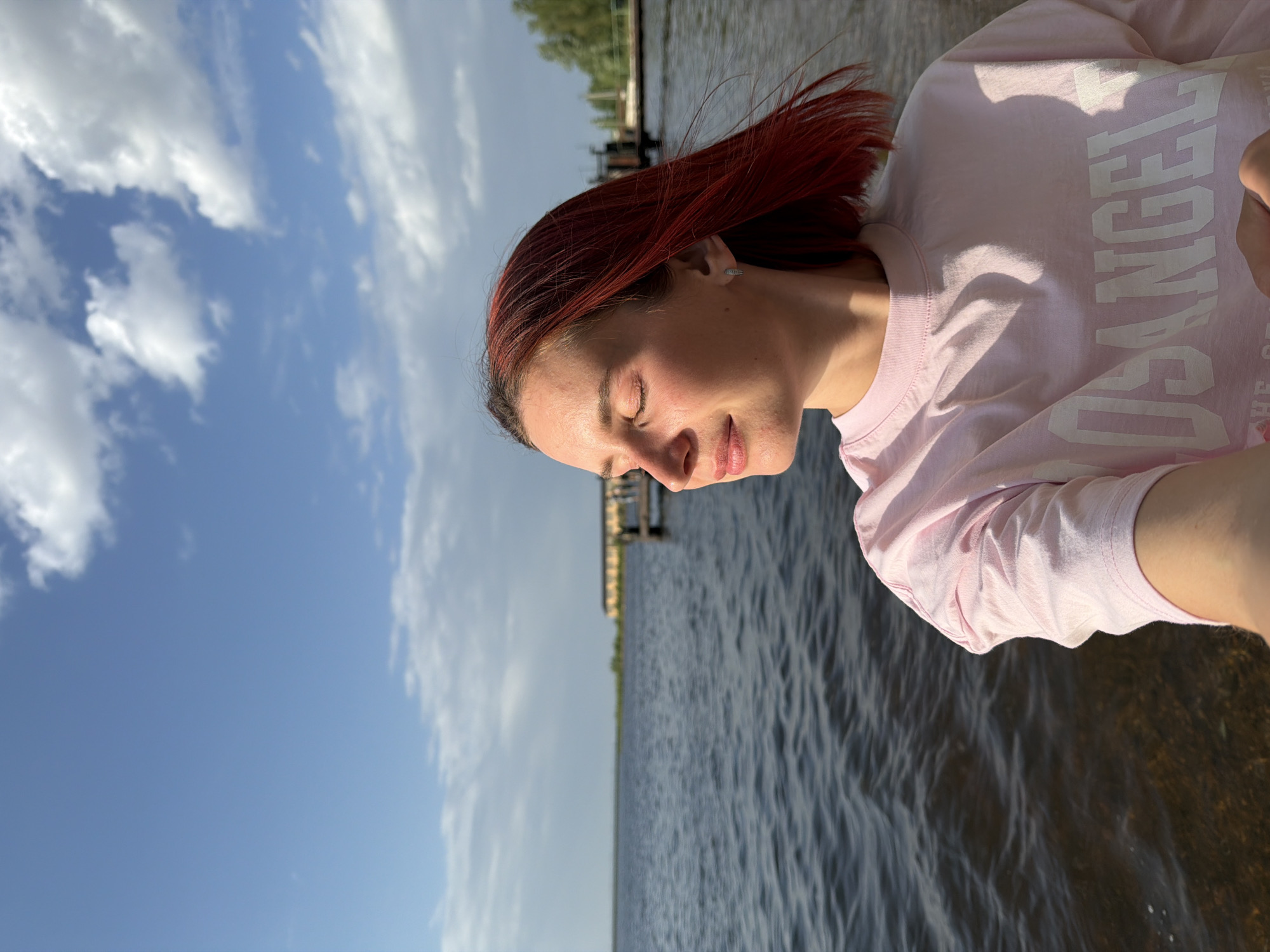 A photograph captured in Chaikovsky, Russia shows a woman with red hair standing outdoors with her eyes closed. She is wearing a pink t-shirt and is positioned in front of a body of water, possibly a lake or river. In the background, there is a pier or dock structure. The sky is blue with scattered clouds, suggesting a bright, sunny day. The woman appears to be enjoying the moment.