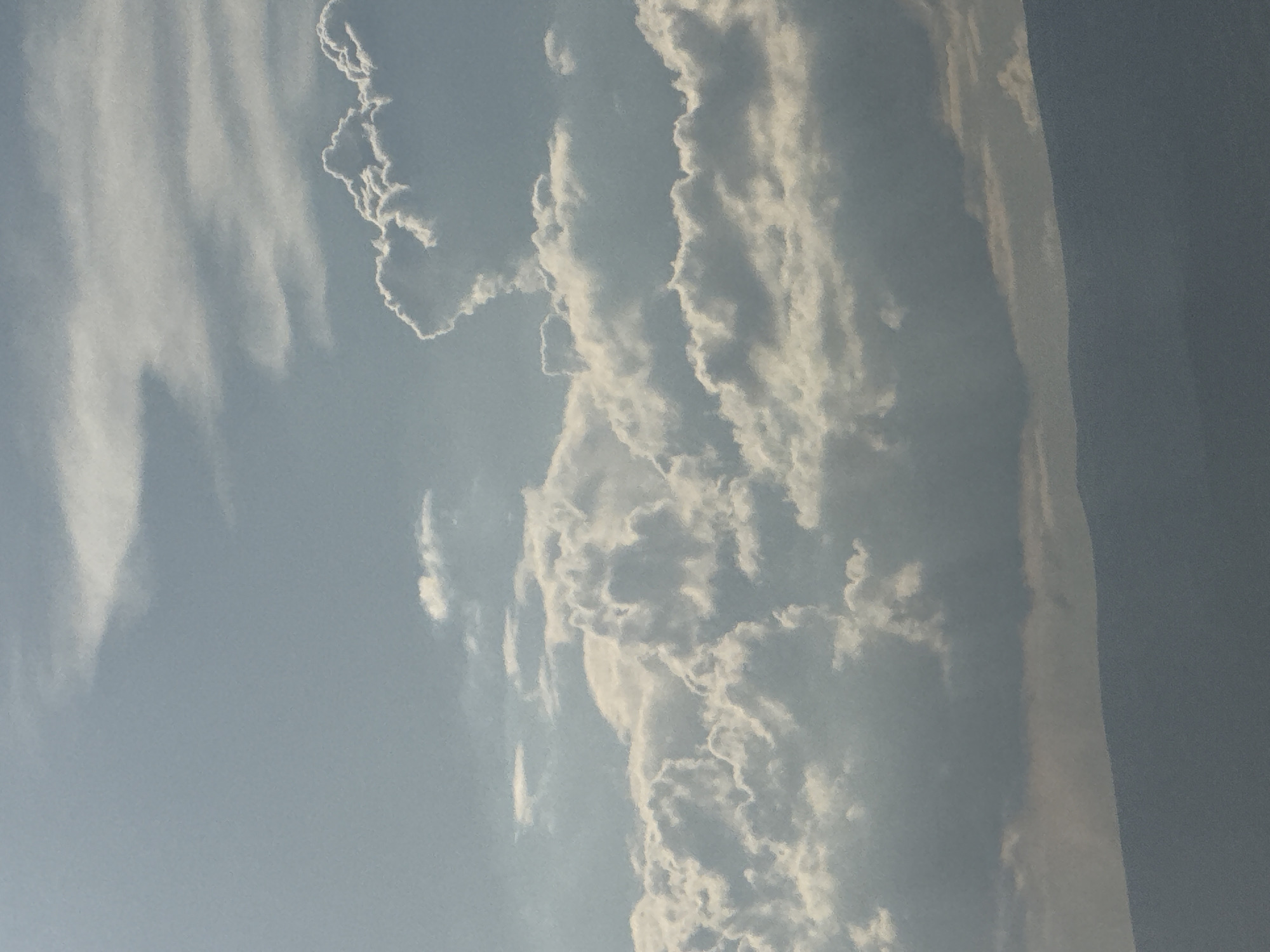 The image displays a wide shot of a hazy, blue sky filled with various cloud formations. In the foreground, the silhouette of a mountain range is visible, a dark blue-gray against the lighter sky. The clouds vary in density and appearance, with some appearing wispy and thin while others are thick and billowy, catching the light and casting shadows. A few scattered, smaller clouds are present in the upper portion of the frame. The overall impression is of a somewhat overcast or diffused lighting condition. No people, specific objects, or discernible actions are present in the image. The scene is devoid of any recognizable landmarks that would specifically indicate the city of Abovyan, Armenia.