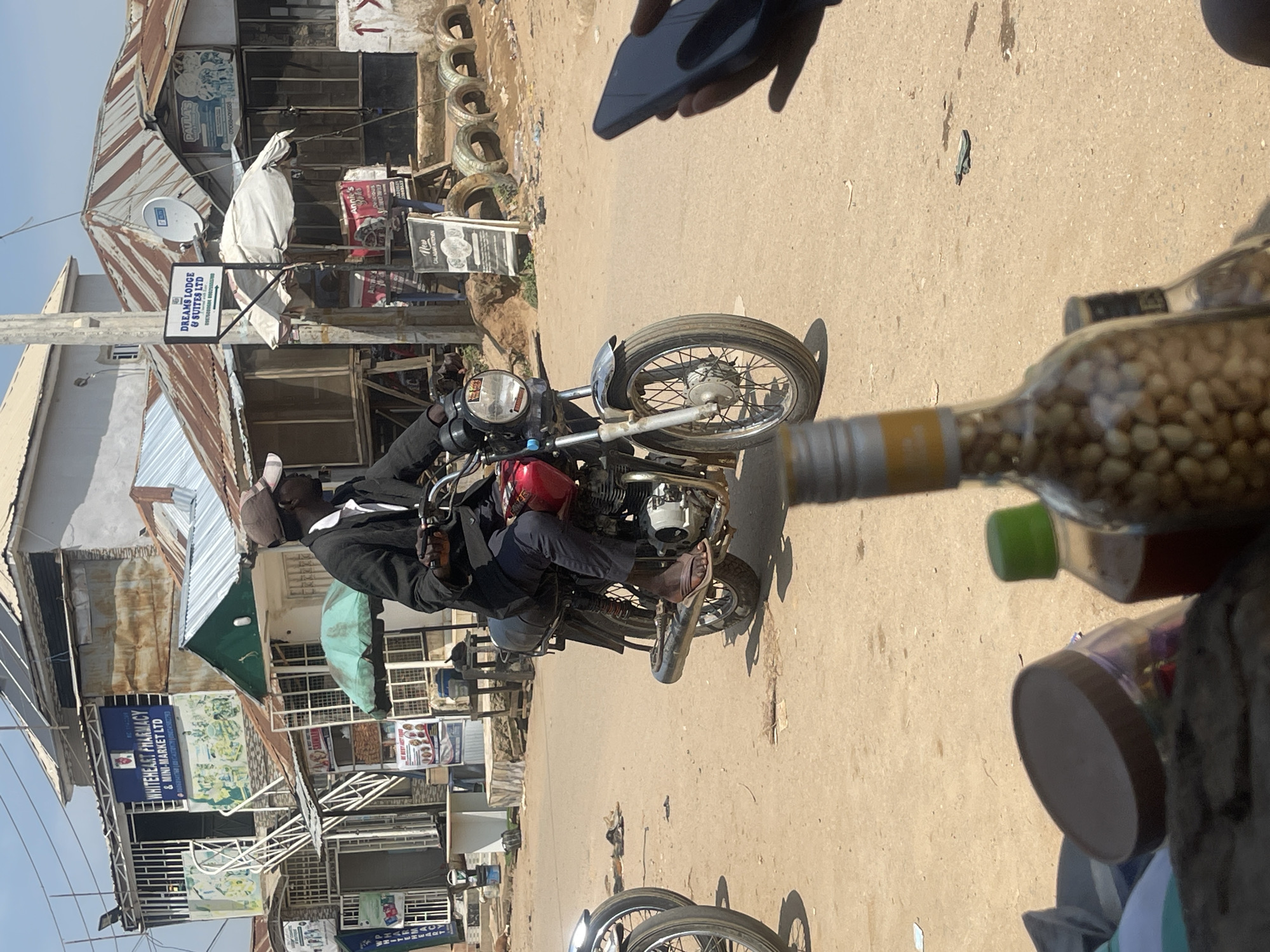 In Abuja, Nigeria, a man wearing a dark jacket and a brown baseball cap rides a red motorcycle with a black seat. He is on a dirt road, and the motorcycle has a chrome exhaust pipe. In the background, there are several buildings with corrugated metal roofs. One building has a sign that reads "WHITEHEART PHARMACY & MINI-MARKET LTD". Another building has a sign that says "DREAMS LODGE & SUITES LTD". There are also some tires stacked on the side of the road and some vegetation. In the foreground, the top of a bottle filled with peanuts is visible, as is a hand holding a dark smartphone.