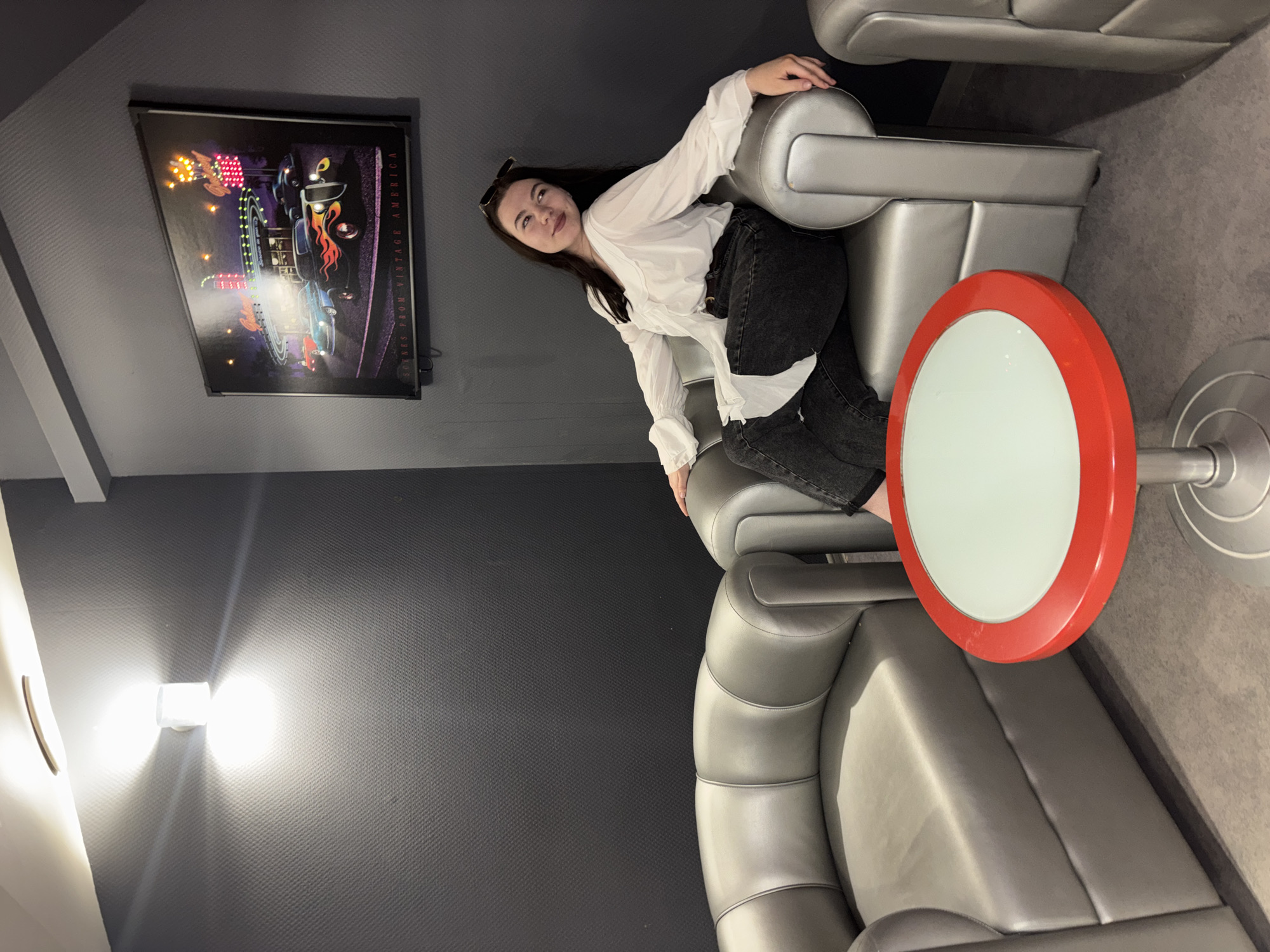A young woman with dark hair is seated on a gray leather chair in what appears to be a lounge area. She is wearing a white shirt and dark jeans. Behind her hangs a framed picture with text on the bottom right side, the picture is mostly black with vibrant colors of yellow, pink, and purple on the left side. A red and white round table is to the right of the chair. The walls are gray. A light fixture is visible on the wall in the lower left of the frame. The scene is likely in a commercial or public space. The location is Perm, Russia.
