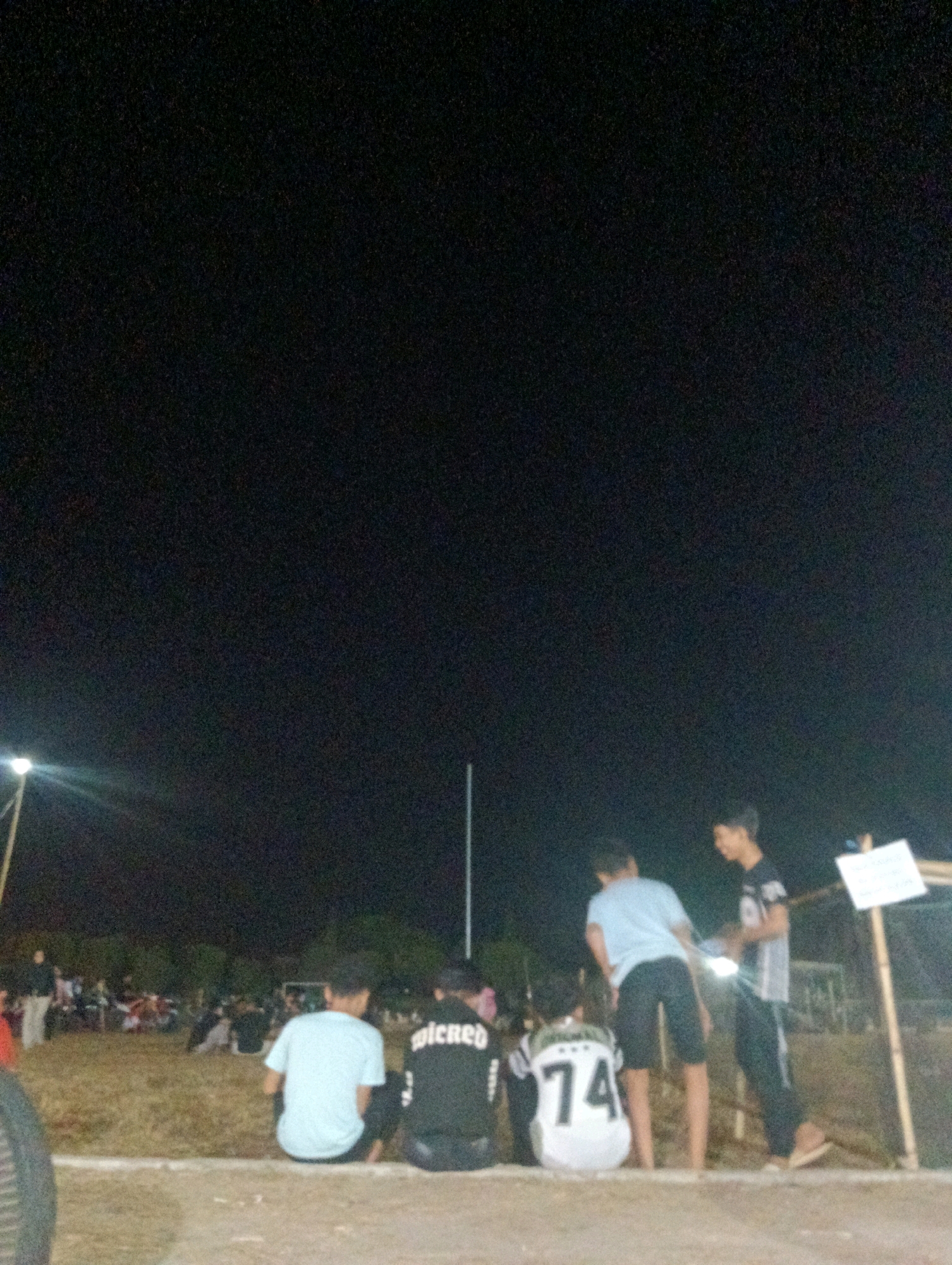 In the city of Pematang Jaya, Indonesia, at night, a group of young males are seated and standing near a field. The setting appears to be a casual gathering, possibly at an outdoor event or sports field, with a dark, unlit background suggesting the absence of ambient city lights. The foreground shows a group of four individuals. Two are seated facing away from the camera, one wearing a light blue t-shirt and dark shorts, and another in a black jacket with white lettering. Another young male is seated between them, facing away, wearing a white jersey with the number "74" prominently displayed in black. The fourth individual is standing, facing right, wearing a black and white t-shirt and dark pants, interacting with someone who is partially visible. In the mid-ground, a crowd of people is visible, some seated and others standing, with what appears to be tents or structures in the background. A tall, thin pole is visible to the right of the standing individuals, with a white sign attached to it. To the left, a brightly lit pole with a light fixture is casting illumination on the scene. The ground in the foreground is a dirt or gravel surface.