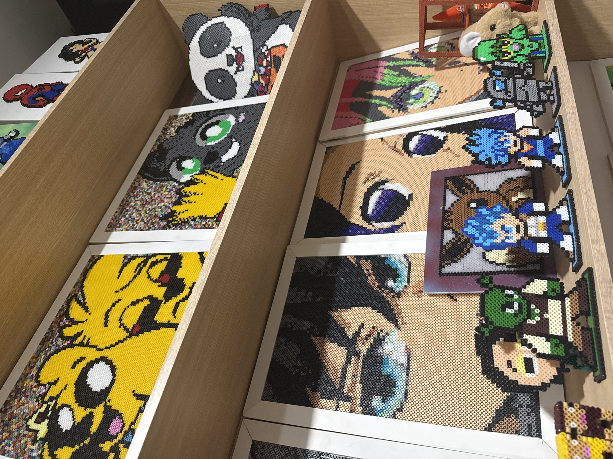 The image shows a display of artwork made from Perler beads, organized in a wooden shelf. The artwork depicts various cartoon and anime characters. From left to right, the shelves contain: a panda head, a black and gray character with green eyes and yellow hair, a yellow character with black features, a portrait of a character with blue eyes, portraits of characters with blue eyes, and pixel art figures of various characters. A stuffed animal toy sits on the top shelf. The environment is indoors, likely a store or gallery. The location is Puebla, Mexico.