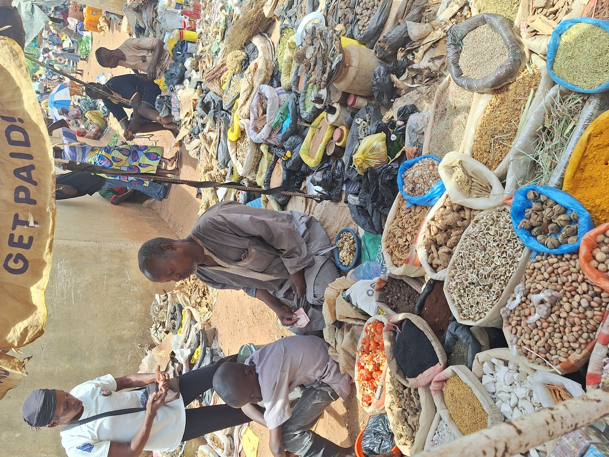 The image shows a market scene in Kakuri Gwari, Nigeria. A man in a grey robe is handing a banknote to a woman in a white t-shirt and black pants. A child in a light-colored shirt is crouching beside them. They are surrounded by bags and containers of various spices, nuts, and dried goods displayed for sale. Other people are visible in the background, along with various merchandise and market infrastructure. The location appears to be an outdoor market with a diverse array of items for sale.