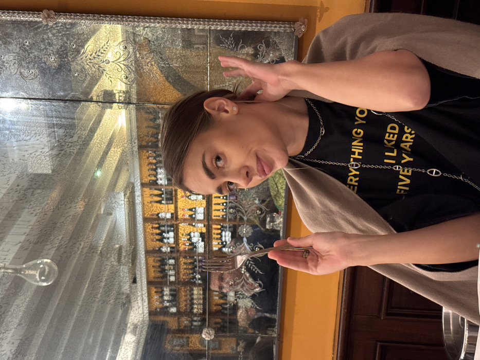 A young adult female is depicted seated at a table inside a restaurant or bar in Venice, Italy. She has brown hair pulled back, a light complexion, and is wearing small stud earrings and a long, thin silver earring threader. She wears a black t-shirt with yellow text reading "EVERYTHING YOU LIKED FIVE YEARS" and two silver chain necklaces with oval links. A light brown shawl is draped over her shoulders. She holds a silver dining fork upright in her right hand, near her face, and her left hand is raised near her left ear. A silver ring is visible on her right ring finger. She looks directly at the camera with a slight smile. Behind her, a large, ornate, aged mirror with decorative etched patterns reflects a bar area. In the reflection, a man with dark hair and a potential nose piercing is visible behind the fork, wearing a dark top with light-colored embellishment. The bar shelves are dark with gold accents and are stocked with numerous bottles. The immediate environment includes a bright orange wall above a dark wood paneled section. A clear glass, tear-drop shaped light fixture hangs from the ceiling, visible in the mirror's reflection.