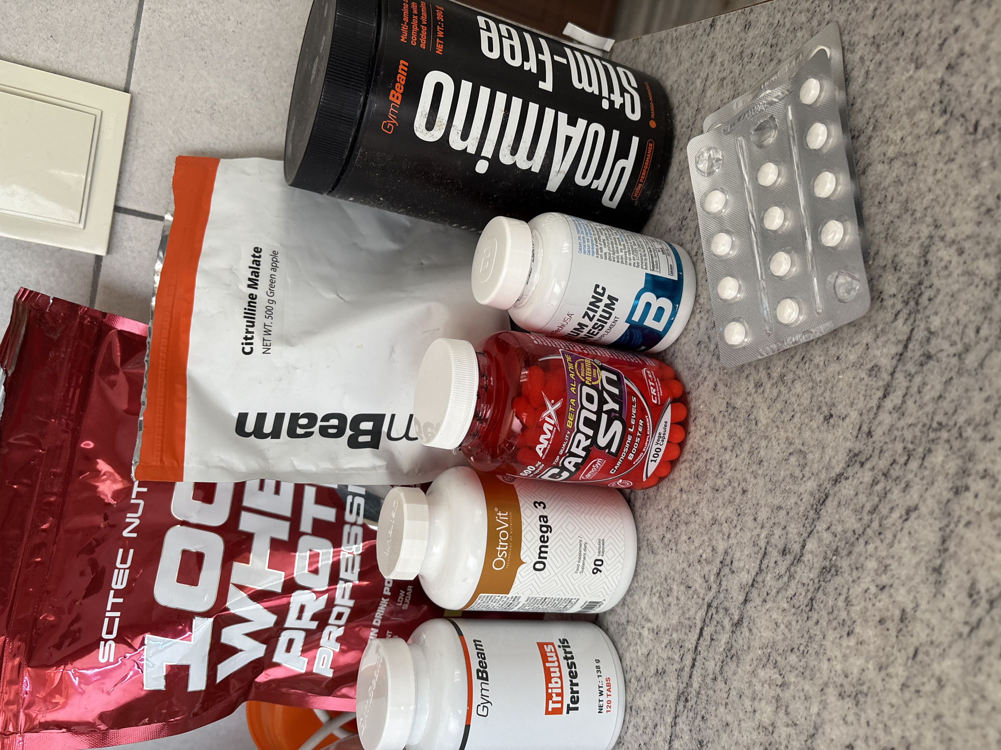 The image shows several dietary supplements and protein products on a mottled gray surface. From left to right, there is a red and white bag of "SCITEC NUTRITION" "100% Whey Protein Professional," a white and orange bag of "GymBeam Citrulline Malate," a black container of "GymBeam ProAmino Stim-Free," a blister pack of white pills, a white bottle of "TechUSA UM ZINC MAGNESIUM," a red bottle of "AMIX CarnoSyn" capsules, a white bottle of "OstroVit Omega 3" capsules, and a white bottle of "GymBeam Tribulus Terrestris" tablets.
There is no indication of the city of Sofia, Bulgaria, or any people or actions in the image.