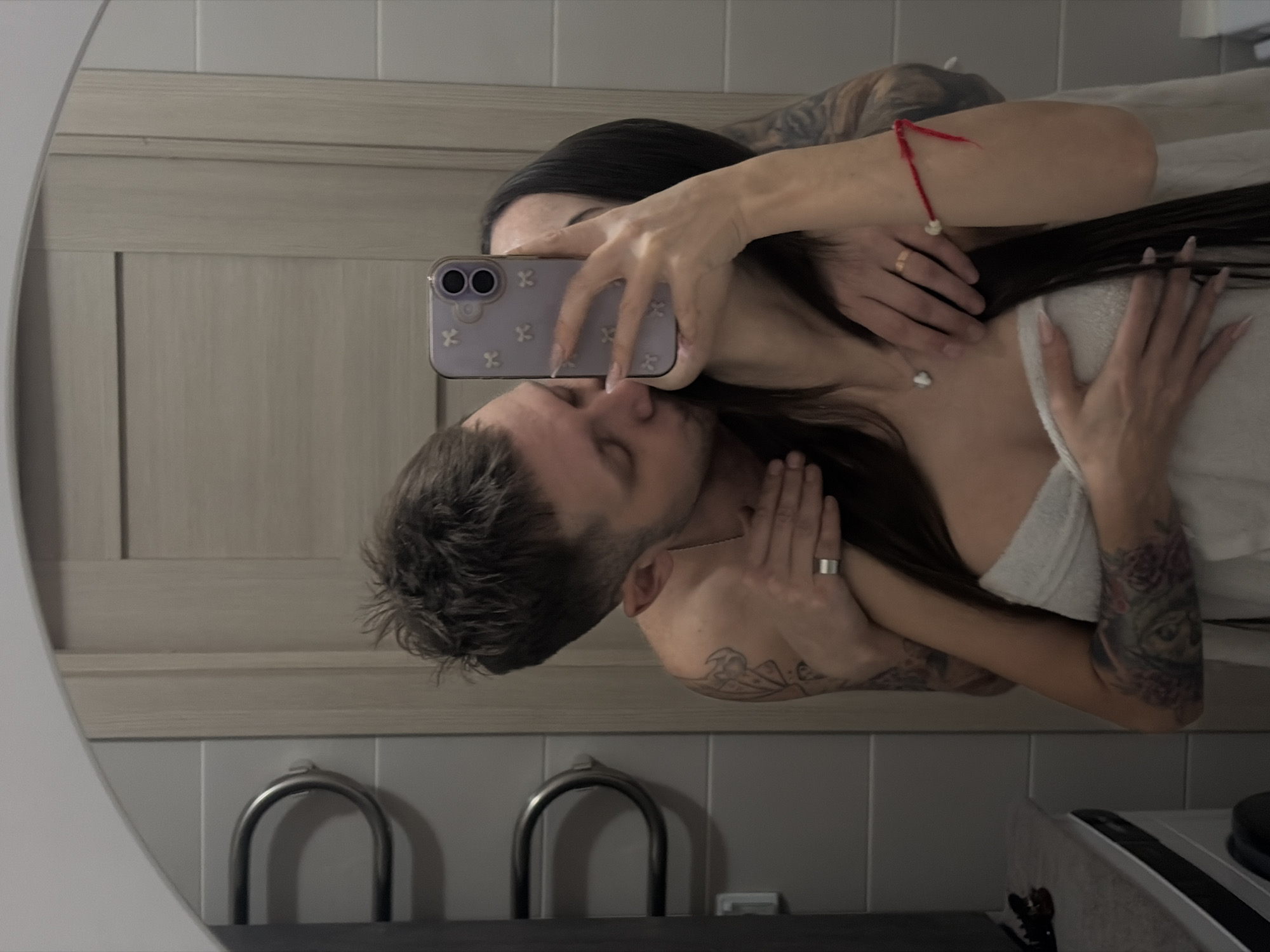 A reflection in a bathroom mirror depicts a couple embracing and kissing. The woman, with long black hair, is holding a purple cell phone with a floral design, ostensibly to capture the scene. Her nails are long and manicured. She is wearing a white covering, and a necklace. Her left arm is adorned with a red bracelet, while her left hand bears a gold ring. The man has short hair and visible tattoos on his neck and arms. The background reveals elements of the bathroom interior including towel holders, a countertop, and tiled walls. The location and city are not discernible from the image.