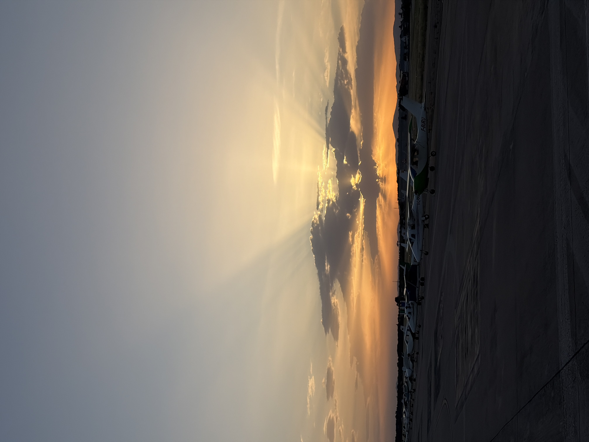 The image shows a row of light aircraft parked on an apron or taxiway, likely at an airport. The aircraft closest to the viewer has the registration "F-BURU" visible on its tail. The background features a sky with dramatic crepuscular rays shining through clouds, suggesting either sunrise or sunset. There is a hint of buildings in the far background. Although the image does not provide explicit details, the mention of the city of Madrid, Spain, suggests that this is likely a view of a general aviation area at Madrid-Cuatro Vientos Airport (LECU), or perhaps another smaller airfield nearby if the location information is assumed to be more approximate. There are no people visible in the image.