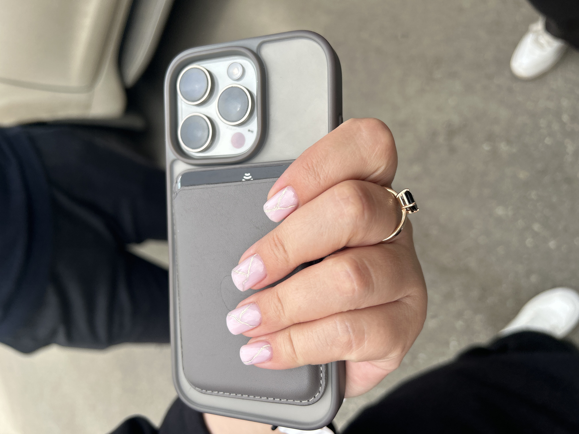 A person's hand, adorned with light pink nail polish and gold geometric accents, is holding a grey phone case. The case features a camera array with multiple lenses. A ring with a dark, rectangular stone is visible on the ring finger. In the background, a person wearing black pants and white sneakers is partially visible, as is a portion of a light-colored vehicle. The ground appears to be paved. The image does not contain any elements that would indicate it was taken in the city of Perm, Russia.
