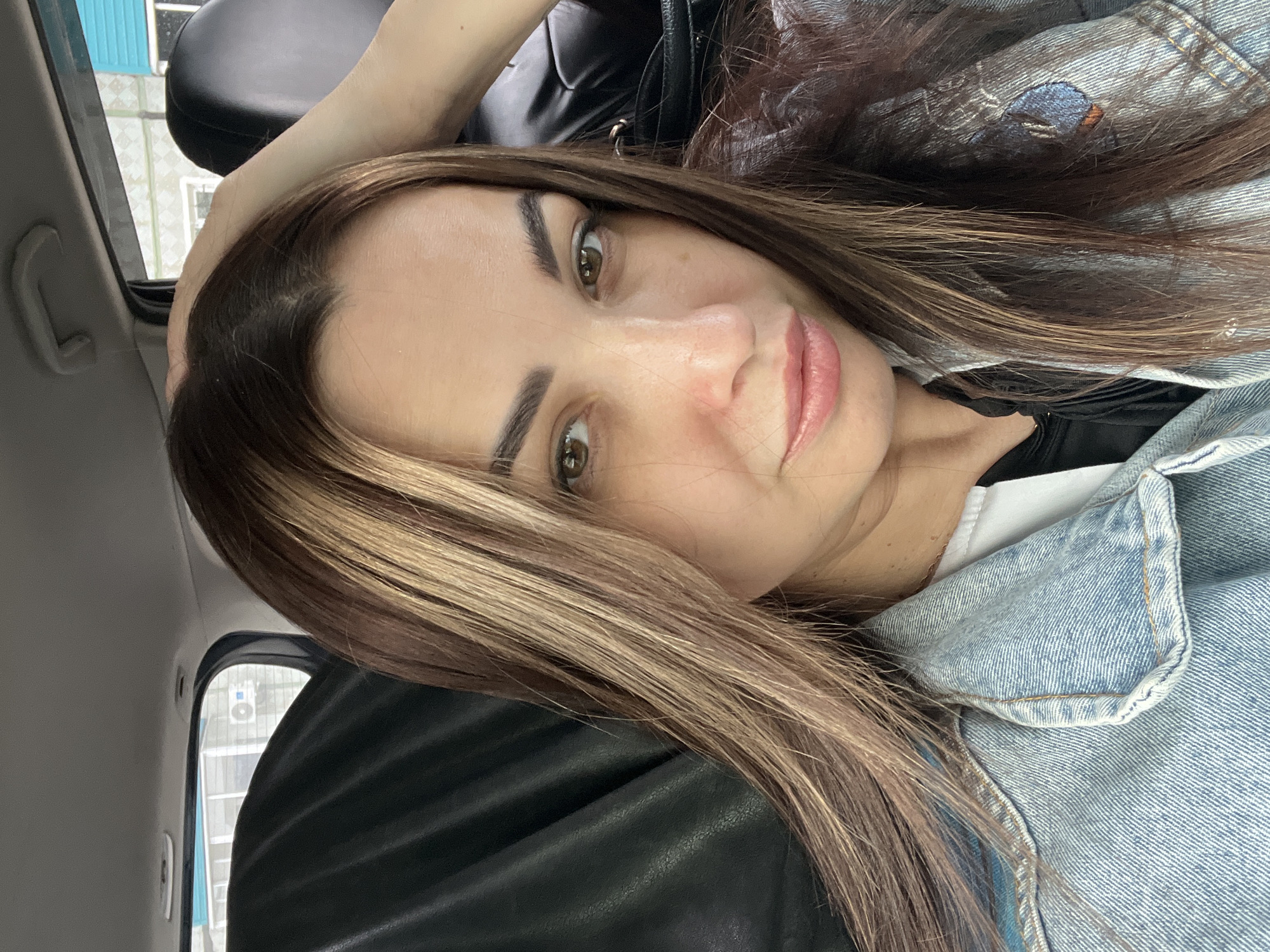 The image is a selfie of a woman with brown hair and hazel eyes, possibly taken inside a car. She is wearing a denim jacket. A black car seat is visible behind her head. A window is on the left side of the frame. The location is unknown.