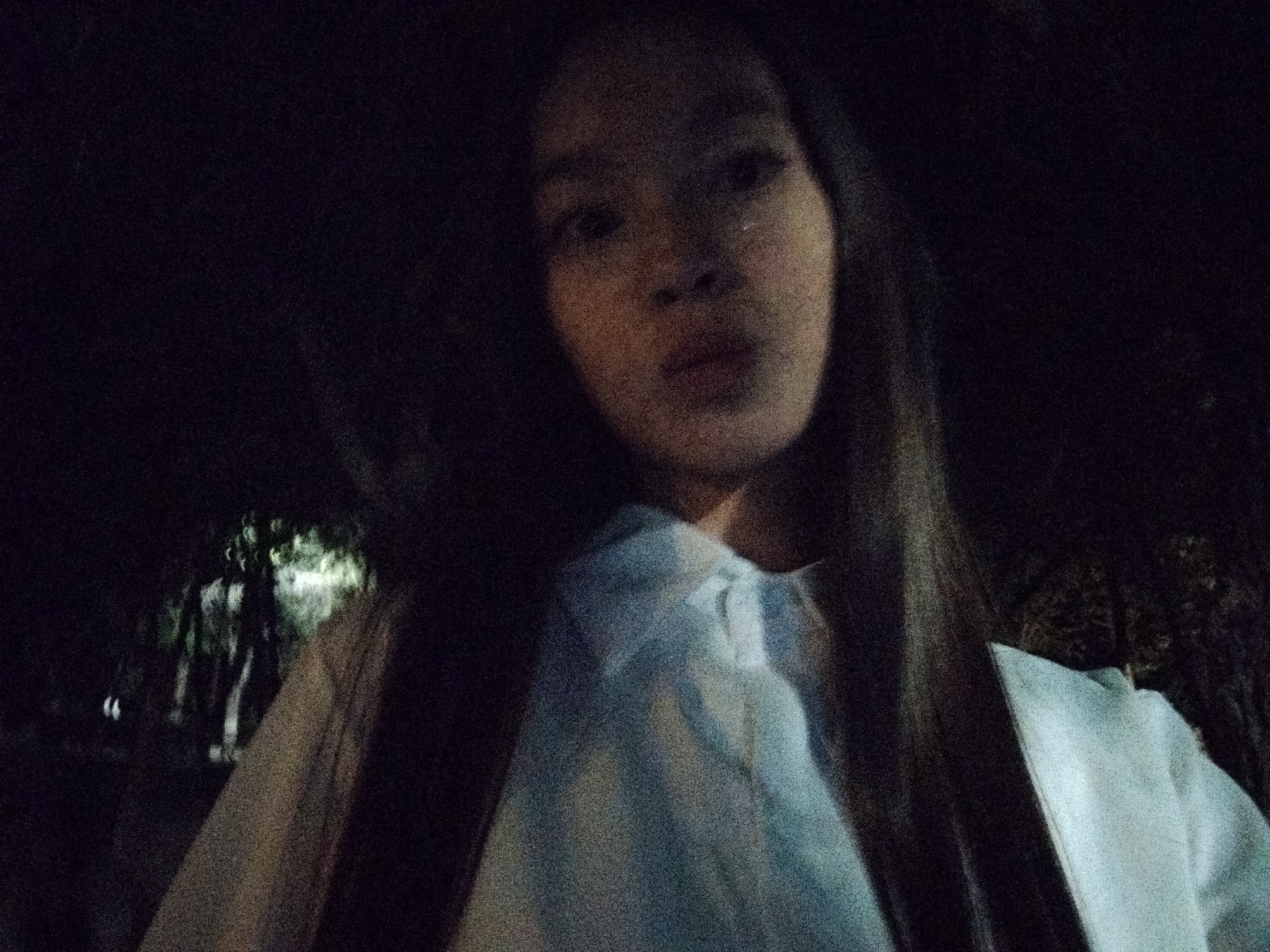The image is a poorly lit selfie taken at night, likely outdoors in a natural or semi-natural environment. A young female with long, dark hair is the central subject, her face partially visible and slightly out of focus. She is wearing a light-colored garment, possibly a shirt or jacket. The background is predominantly dark, with some faint suggestions of trees or foliage illuminated by an unseen light source. The overall impression is of a candid, low-light photograph. There are no identifiable landmarks or structures that would definitively place this image in Bishkek, Kyrgyzstan.