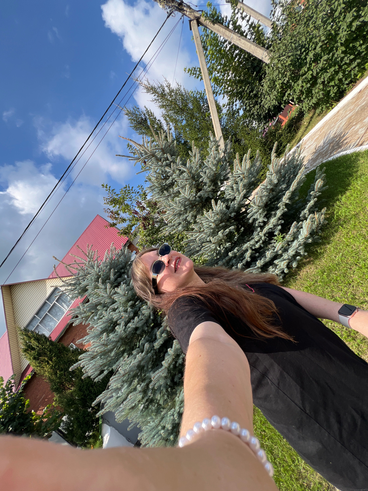 In Noviy, Russia, a young woman with long brown hair and sunglasses takes a selfie outdoors. She is wearing a black t-shirt and a pearl bracelet on her left wrist. She also has a smartwatch on her right wrist. Behind her, a blue spruce tree and a red-roofed house are visible. The sky is blue with some white clouds, and power lines are strung overhead. The scene appears to be a residential area with a grassy lawn and a paved walkway.
