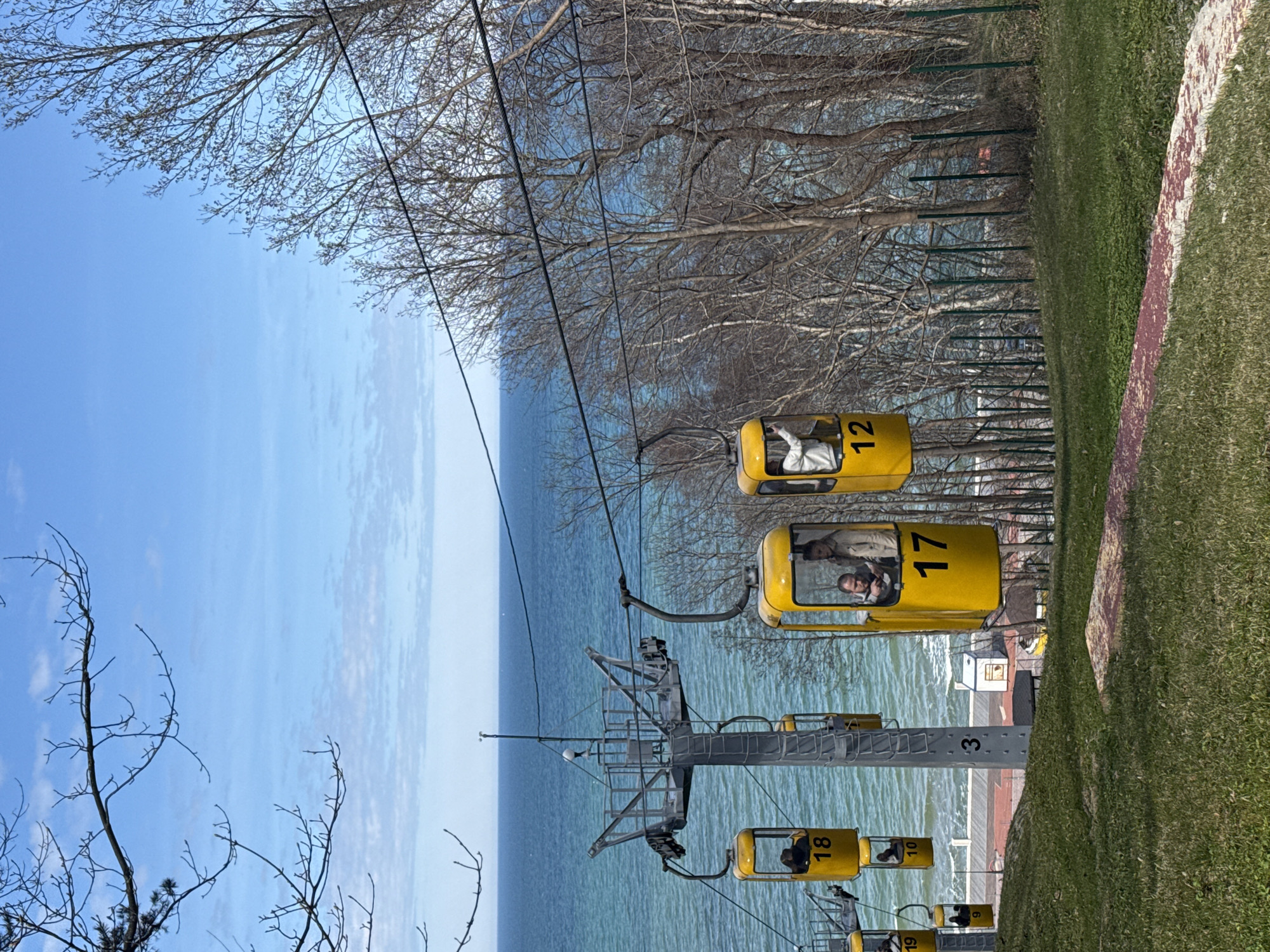 The image depicts a scenic view in Svetlogorsk, Russia, featuring a yellow cable car system against a backdrop of the sea and a partly cloudy sky. The cable cars are numbered 12, 17, and 18, respectively. Passengers are visible inside some of the cars. The structure supporting the cables is a tall metal tower, marked with the number 3. The surroundings show trees with bare branches. Grass and a concrete barrier define the boundary of the area near the cable car base.
