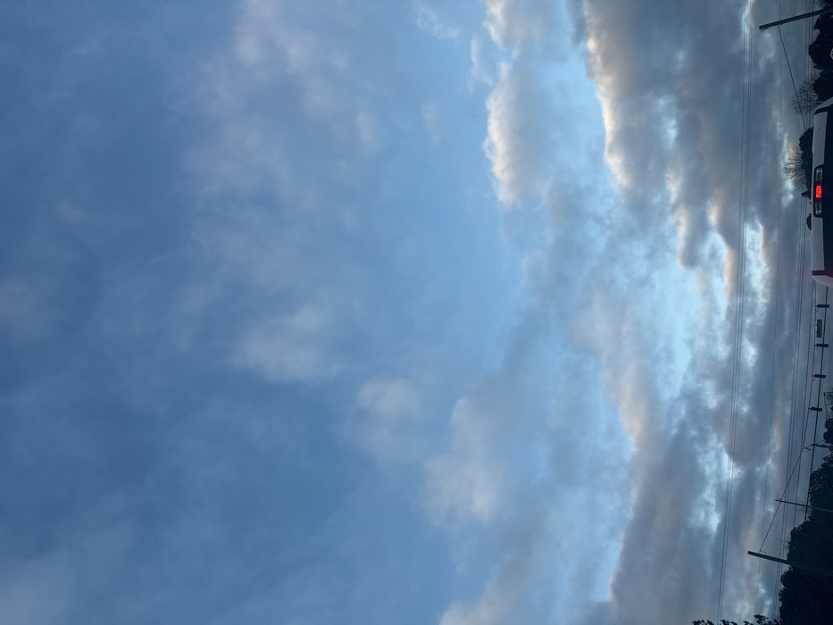 The image displays an outdoor scene in Lawrenceville, United States, focusing on the sky and elements of a street environment. The upper two-thirds of the frame consists of a light blue sky with scattered white and grey cloud formations. Across the mid-ground, multiple dark utility poles and overhead wires are visible, with dark tree lines extending beneath them. In the immediate foreground, the top-rear section of a light-colored pickup truck is partially visible, featuring a central red brake light illuminated on its tailgate. Above the truck, several dark traffic light fixtures are suspended from overhead wires. No individuals are present in the scene, and no discernible actions or interactions are occurring.