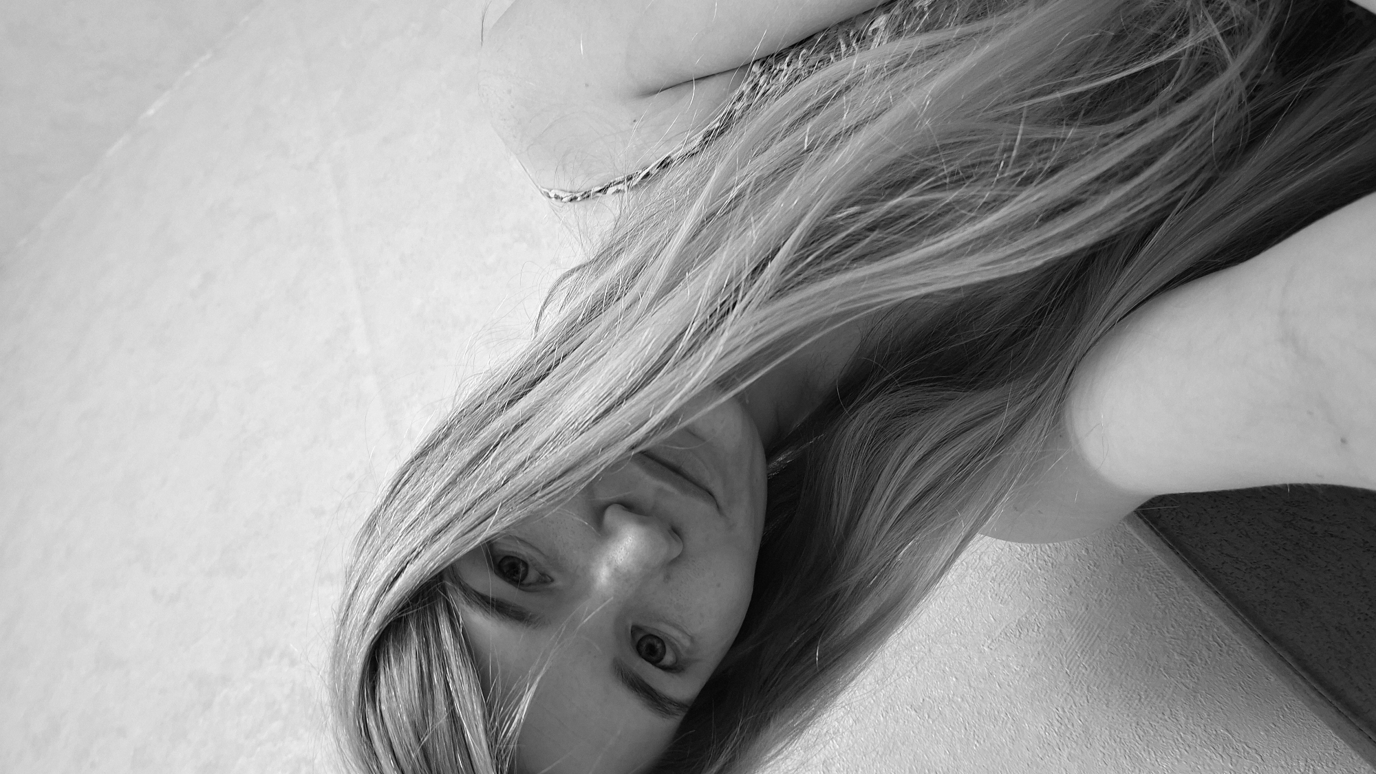 This is a black and white, low-angle, close-up, selfie-style photograph of a young woman with long, blonde hair. The woman's face is partially obscured by her hair and is tilted slightly. Her eyes are open, and she appears to be looking directly at the camera. She has a fair complexion and some freckles are visible on her face. Her hair is long and appears to be straight, with some strands falling across her face. She is wearing a patterned top with a strap visible over her shoulder. The background is a textured, light-colored surface, possibly a wall, and a darker surface which might be furniture or a wall in shadow. The lighting appears to be soft. The image is oriented sideways, suggesting it was taken with the camera held vertically. The location context provided is Большой Букор, Russia, but there are no specific visual cues within the image to confirm this location. No text is visible in the image.