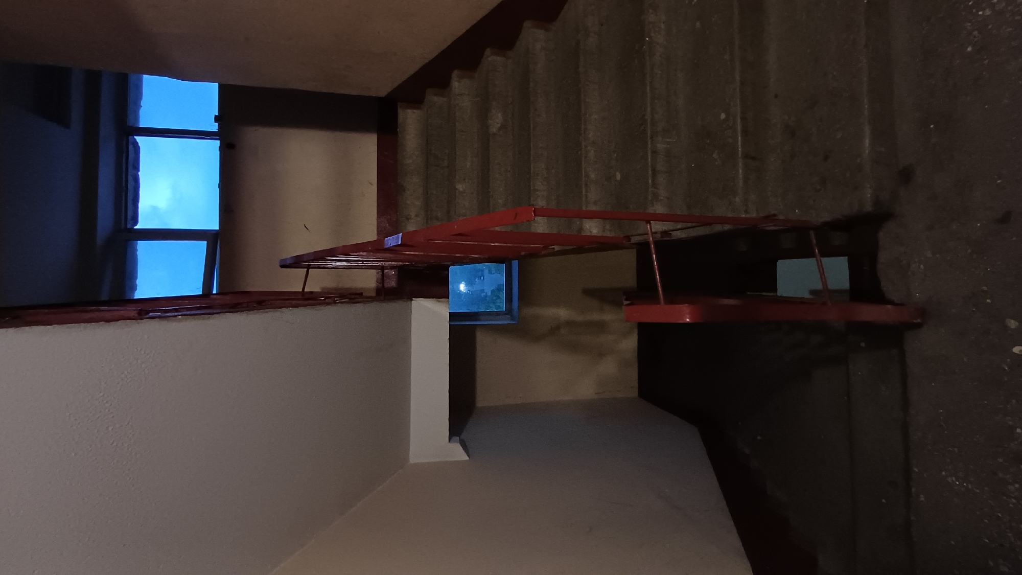 The image shows an interior stairwell, potentially in a multi-story building in Balashikha, Russia. The focus is on the junction between the stairwell landing and the ascending staircase. A window is visible on the landing, providing a view of the sky outside. The stairs are concrete, and a red metal railing runs along the stairs. The walls on the landing are painted white. There are no people visible in the image.