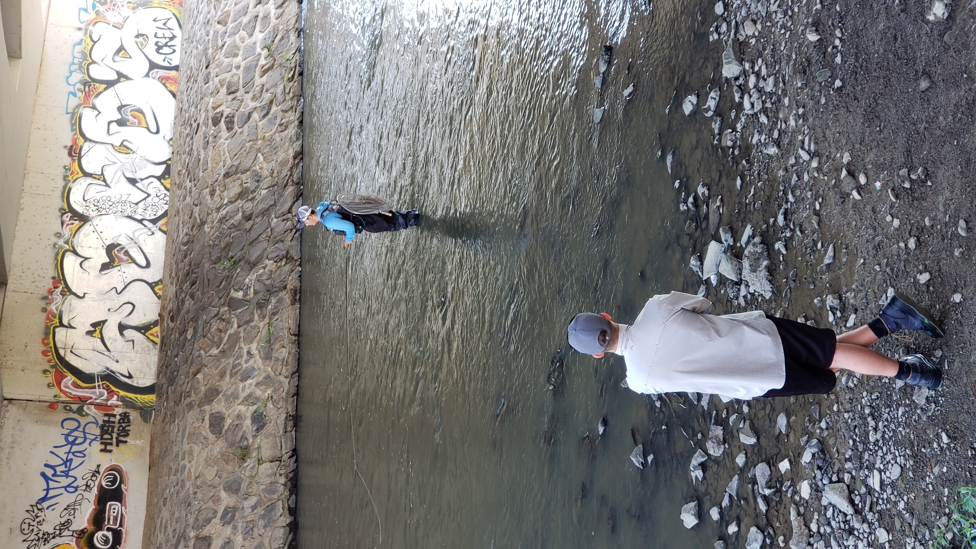 In Prešov, Slovakia, under a concrete bridge adorned with graffiti, two individuals are engaged in fly fishing. The river, reflecting the overcast sky, flows past a rocky embankment. In the foreground, a young person stands on the shore, facing away from the camera, wearing a light-colored shirt, black shorts, socks, and dark sneakers. They appear to be observing the other angler. Further in the river, a second person, dressed in waders, a blue long-sleeved shirt, and a life vest, is actively fishing. This individual holds a fishing rod with a line extending into the water, and a creel is slung over their back. The setting suggests a recreational activity taking place in an urban waterway.