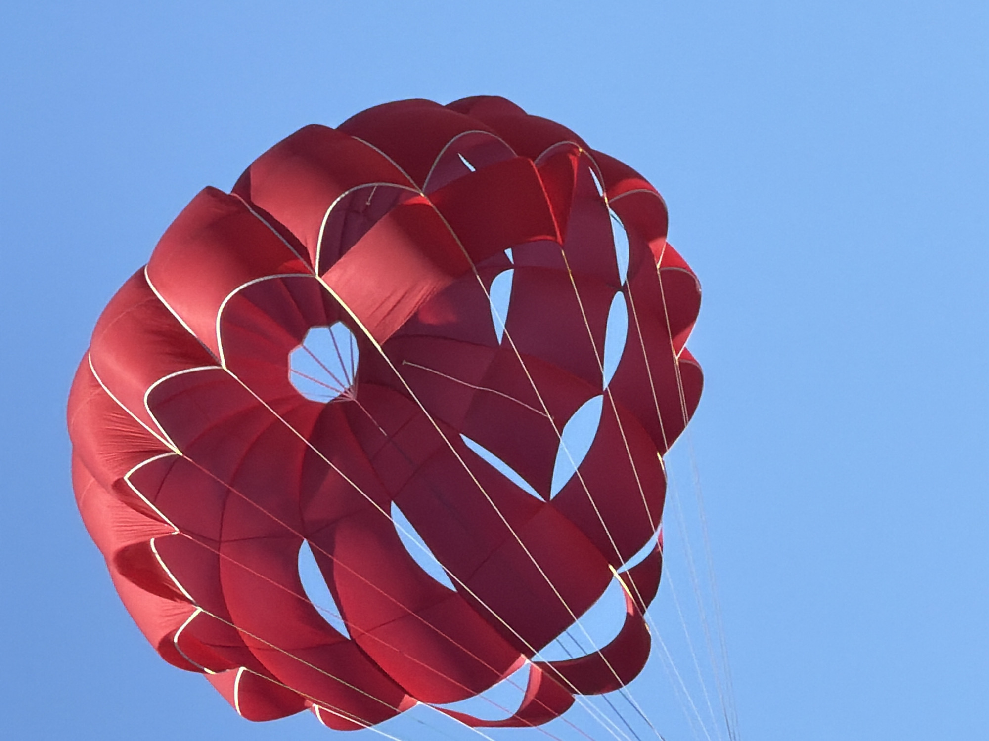 A low-angle shot depicts a red parachute against a clear blue sky, evoking a sense of recreational activity. The parachute is circular with rounded segments edged in gold. Several gaps are visible within the parachute, revealing the sky behind. White strings extend from the bottom of the parachute downwards. The image does not depict any people, other objects, actions, or interactions within a scene located in Bečići, Montenegro.