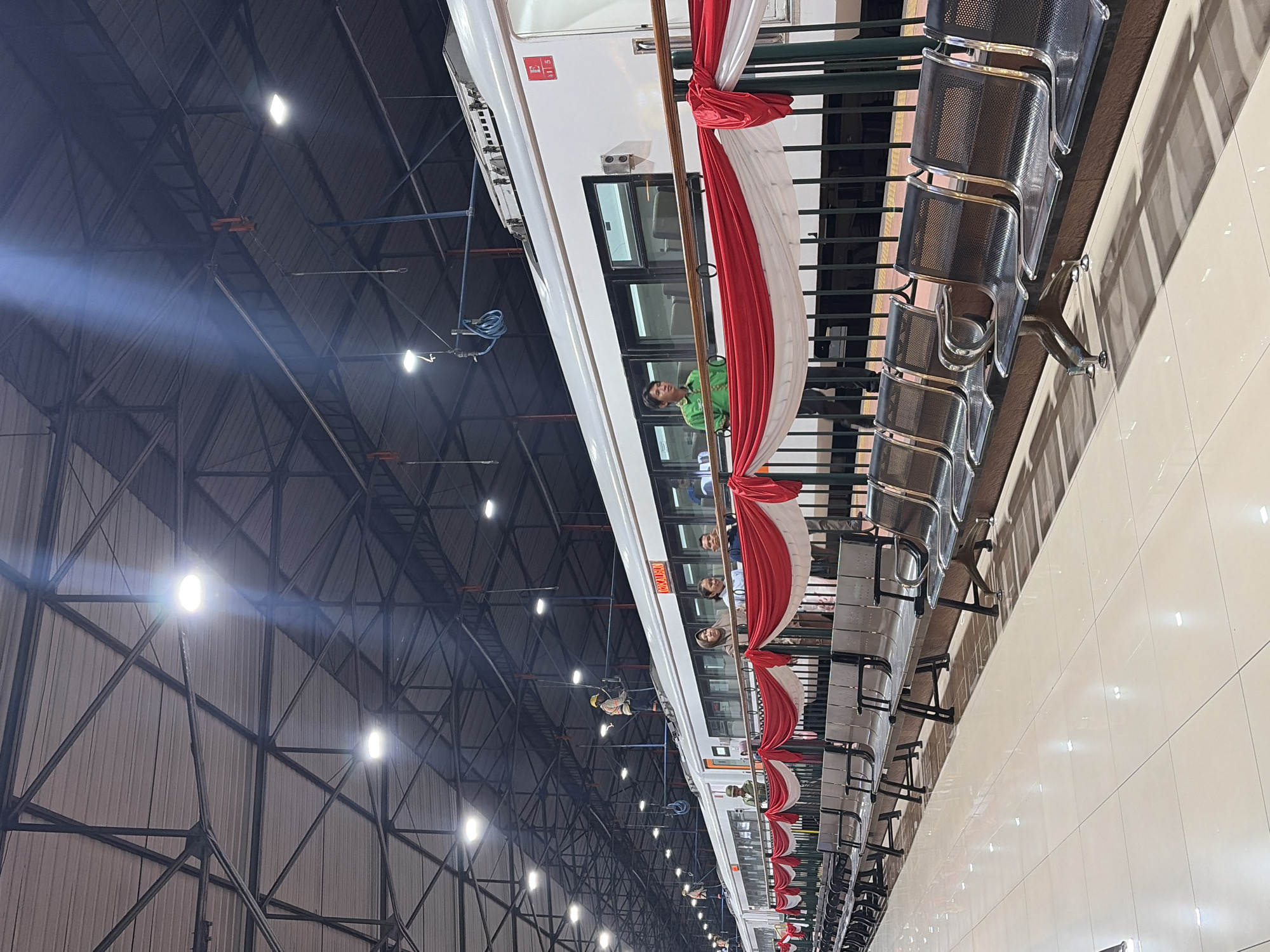 The image shows a train at a station in Semarang, Indonesia. The train is white with red and white banners draped along its side. Several people are visible on the train and on the platform. The platform is lined with empty seats. The station has a large, open roof structure with industrial lighting.