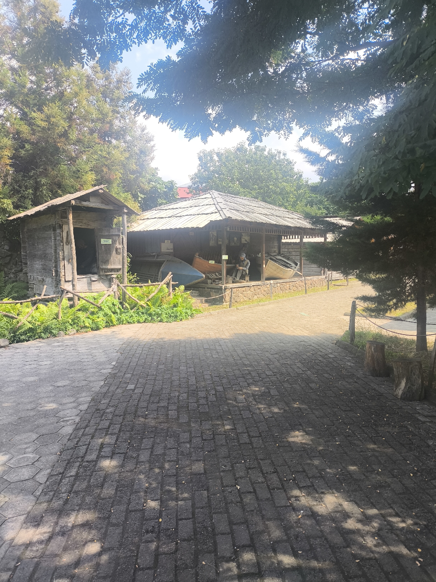 The image shows an outdoor exhibit of what appears to be traditional boats and boat-making structures, possibly located in the city of Batumi, Georgia. In the foreground, a paved walkway made of interlocking gray bricks leads towards the exhibit. The walkway is partially covered in shade from overhead trees. To the left of the walkway is a small, rustic wooden structure with an open doorway, revealing a dark interior. In front of this structure, a low wooden fence encloses a patch of green ferns and foliage.
Further into the scene, a larger, open-air structure with a thatched roof houses several boats. One boat, a long, narrow vessel, is prominently displayed on its side. Another, larger boat is positioned in the background. A mannequin dressed in work clothes is seated inside this structure, seemingly engaged in boat construction. The exhibit is surrounded by lush greenery and trees, creating a natural, somewhat enclosed atmosphere. The overall impression is of a historical or cultural exhibit showcasing maritime traditions.