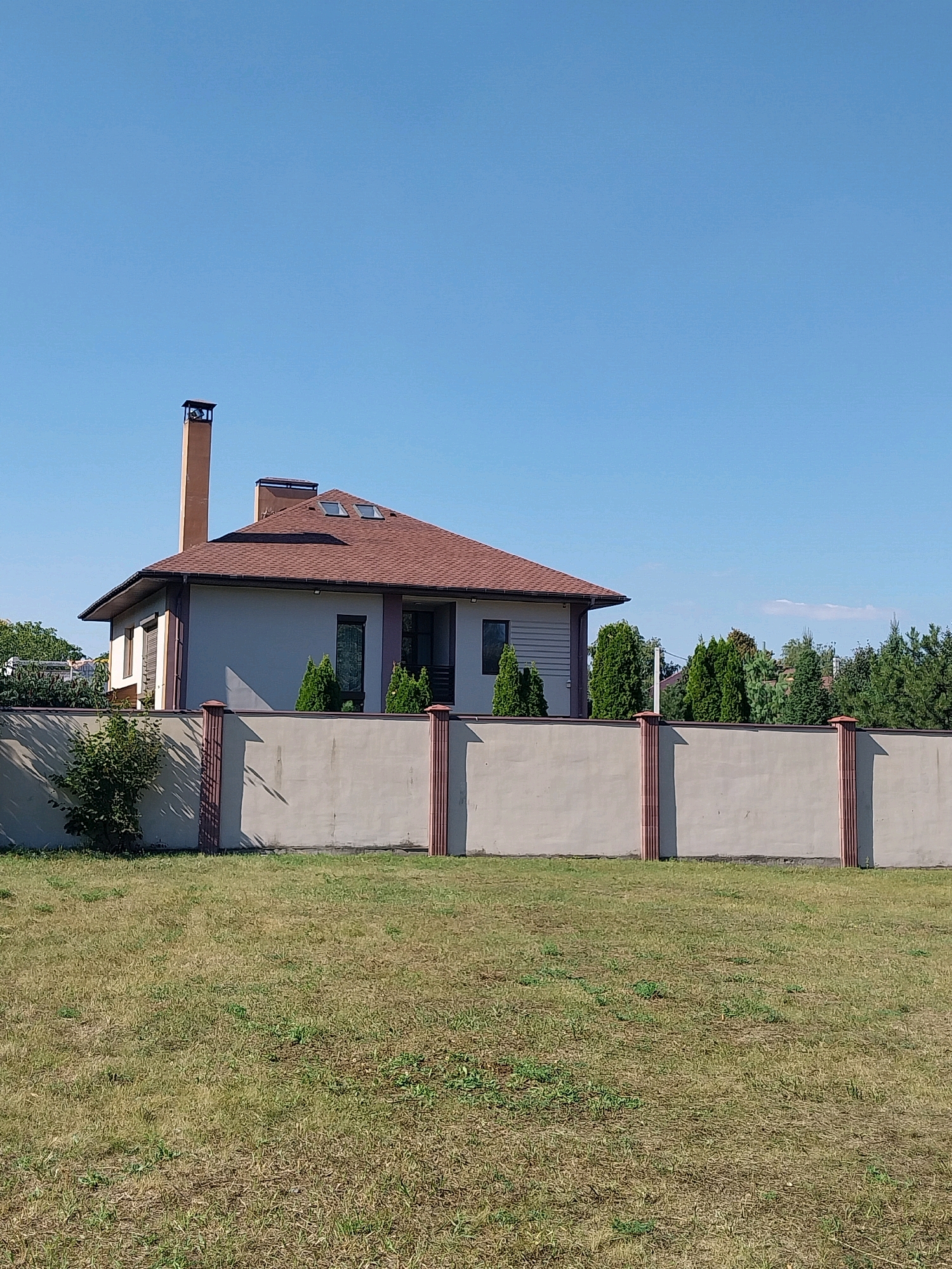 In the city of Doslidne, Ukraine, a modern, two-story house with a brown shingled roof is visible, featuring a tall, brick chimney. The house is partially obscured by a tall, light gray concrete fence with decorative brown pillars. The yard in front of the fence is covered in dry, yellow-green grass, with a few scattered green weeds. Behind the fence, several well-maintained evergreen trees are planted in front of the house. The sky is clear and blue.