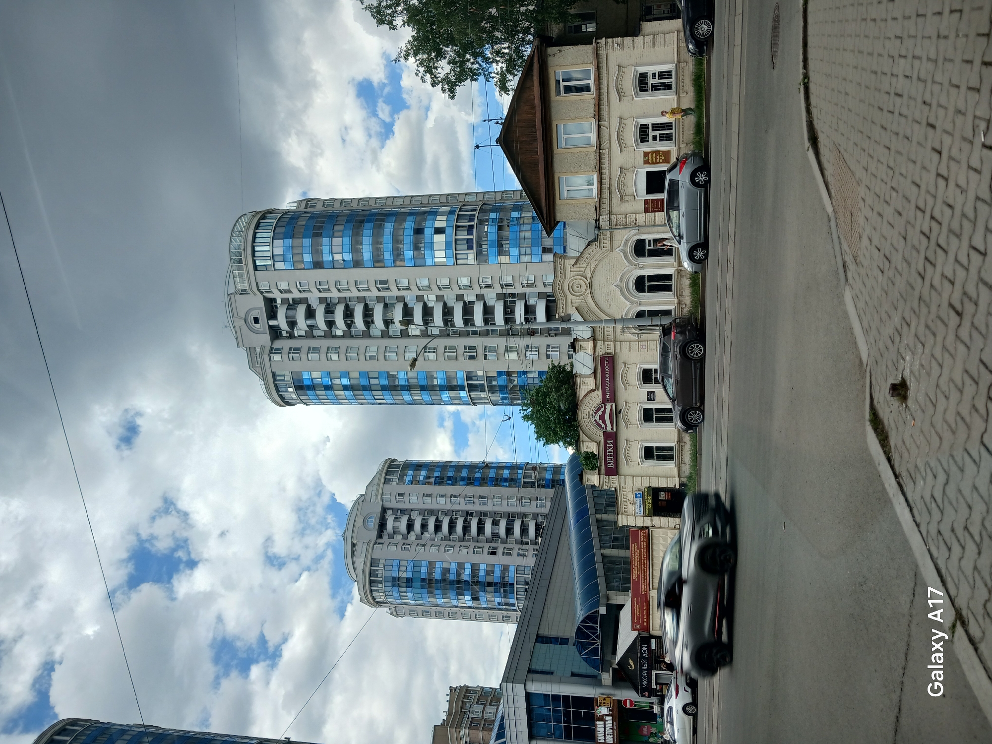 Here's a detailed description of the image:
**Setting:** The image shows a street scene in Yekaterinburg, Russia. The perspective is from the roadside looking towards buildings.
**Buildings:**
* A modern cylindrical high-rise with blue reflective windows and white balconies dominates the left side. There are at least two of these towers visible, one partially obscured.
* An older, two-story building with decorative architecture sits beside the modern towers. It has a beige facade, a dark wooden roof, and arched windows. This building houses businesses, with signs reading "ВЕНКИ" and "ПРИНАДЛЕЖНОСТИ".
**Street and Vehicles:**
* A paved road runs along the right side of the image.
* Several cars are parked or moving along the road. A black car is blurring due to its motion.
* A sidewalk made of cobblestones is visible on the right edge of the frame.
**Sky:**
* The sky is cloudy with a mix of dark storm clouds and patches of blue sky.
**Signage:**
* Various signs in Russian are attached to the older building, including those for "ИКОРНЫЙ ДОМ" (caviar house) and "БОЛЬШОЙ ЦВЕТОЧНЫЙ" (large flower shop).
**Text Overlay:**
* The text "Galaxy A17" is displayed in the lower right corner.