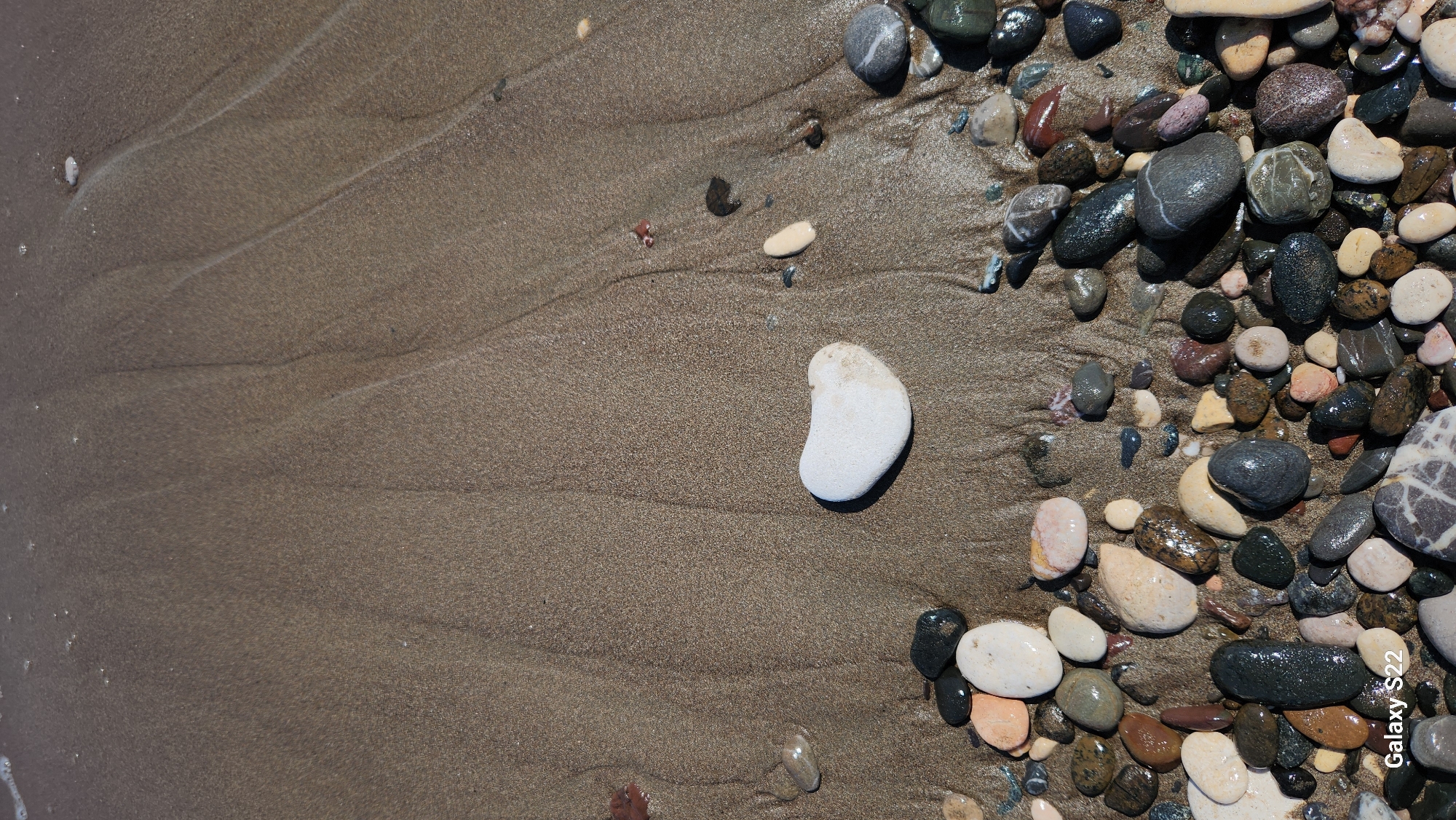 The image displays a high-angle, close-up view of a pebbly and sandy beach at the water's edge in Erdemli, Turkey. The upper half of the frame consists of light brown, damp sand exhibiting subtle ripple patterns from receding water. Minimal white foam is visible along the top edge, indicating the maximum reach of the tide. Below this, the sand transitions to a darker, wetter consistency.
A single, smooth, white stone, irregularly shaped and approximately heart-like, rests centrally on the damp sand in the lower-middle section; its right side shows a slight tan stain. The bottom third of the image is densely populated with various-sized, water-saturated pebbles. These pebbles are diverse in color, including shades of black, dark grey, white, cream, tan, and reddish-brown, with many appearing shiny due to wetness. Some pebbles exhibit distinct patterns, such as a grey stone with white stripes located in the bottom right corner. No human figures or activities are present. The text "Galaxy S22" is visible in white lettering in the bottom-left corner of the image.