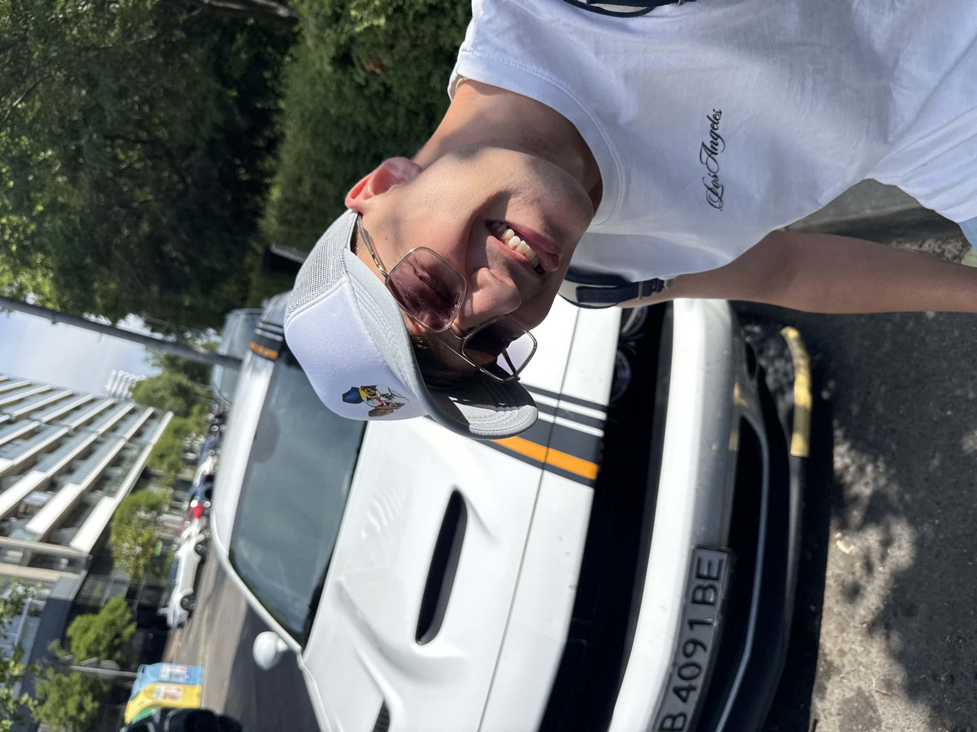 A young man with a white mesh-back cap and sunglasses smiles at the camera, leaning in from the right side of the frame. He wears a white t-shirt with "Los Angeles" printed on it in black script. Behind him, a white Dodge Challenger is parked on a paved street, with its front bumper and hood visible. The car has a black grille and racing stripes in orange and dark gray running down the center of the hood and bumper. The license plate reads "B 4091 BE". The scene appears to be in an urban environment, with a modern apartment building and trees lining the street. The lighting suggests it is daytime. The location is identified as Burgas, Bulgaria.