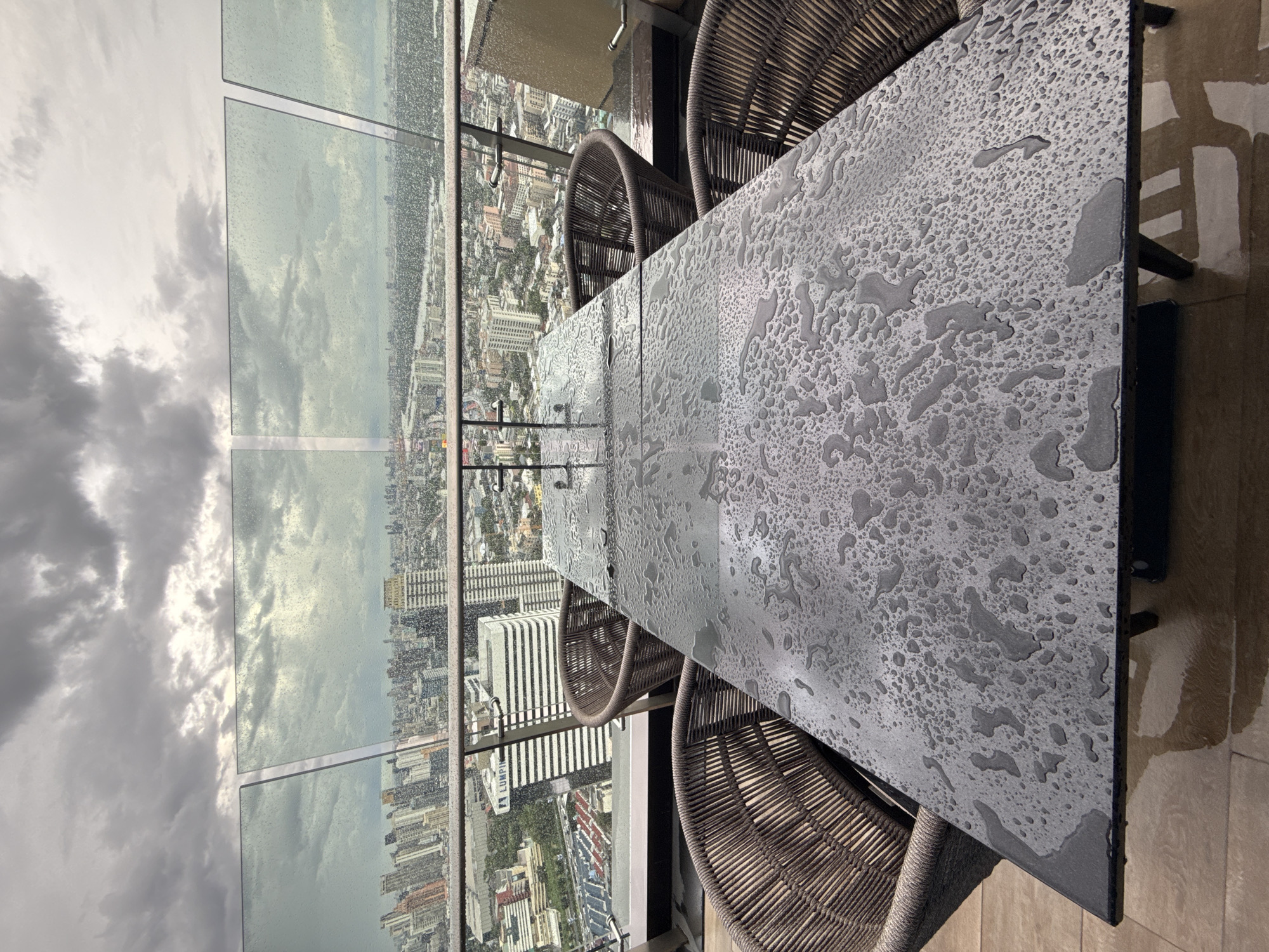 The image depicts a rooftop dining area in Bangkok, Thailand, during a rain shower. A rectangular dark-colored table is covered in numerous water droplets, indicating recent rainfall. Around the table, four woven wicker chairs are visible. Beyond the table and chairs, a glass barrier separates the seating area from the cityscape. The view beyond the barrier reveals the dense urban landscape of Bangkok, with numerous skyscrapers and buildings stretching towards the horizon. The sky is overcast with dark, heavy clouds, and rain streaks are visible on the glass barrier, further emphasizing the rainy weather. The flooring of the rooftop area appears to be made of wooden planks, which are wet and reflective from the rain. The overall scene conveys a sense of quietude and a dramatic urban panorama subdued by the weather.