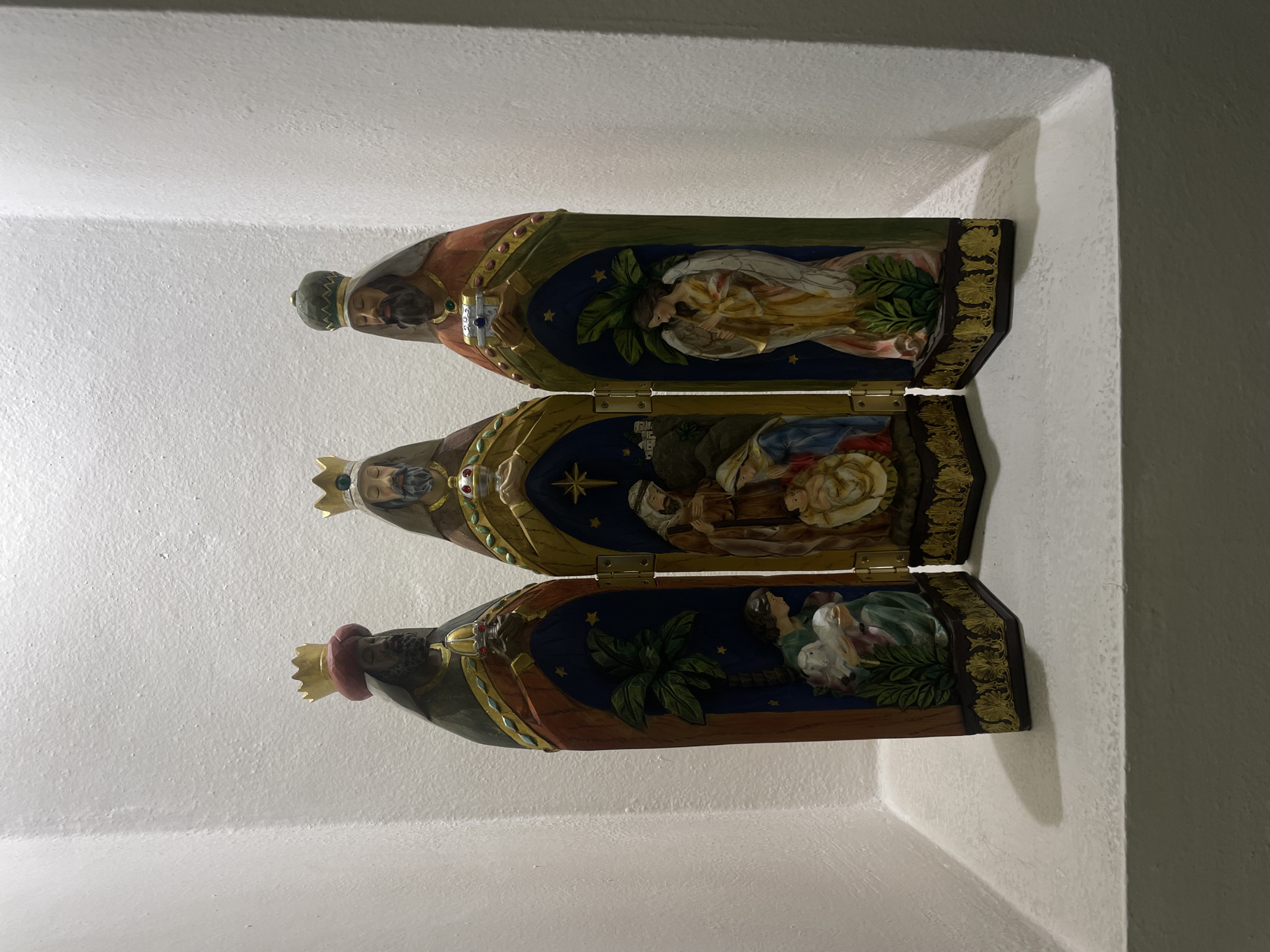 The image shows a three-panel religious triptych of the Three Kings (Three Wise Men) displayed inside a recessed niche in a wall. The triptych is made of wood and features carved figures of the kings on the outside panels. The inside panels depict scenes from the Nativity story, including an angel, the baby Jesus, and Mary and Joseph. The triptych has ornate gold detailing around the edges and is secured with hinges.
The wall and niche are painted white, and the lighting is somewhat dim, suggesting an indoor setting. The image was taken in Oaxaca City, Mexico.