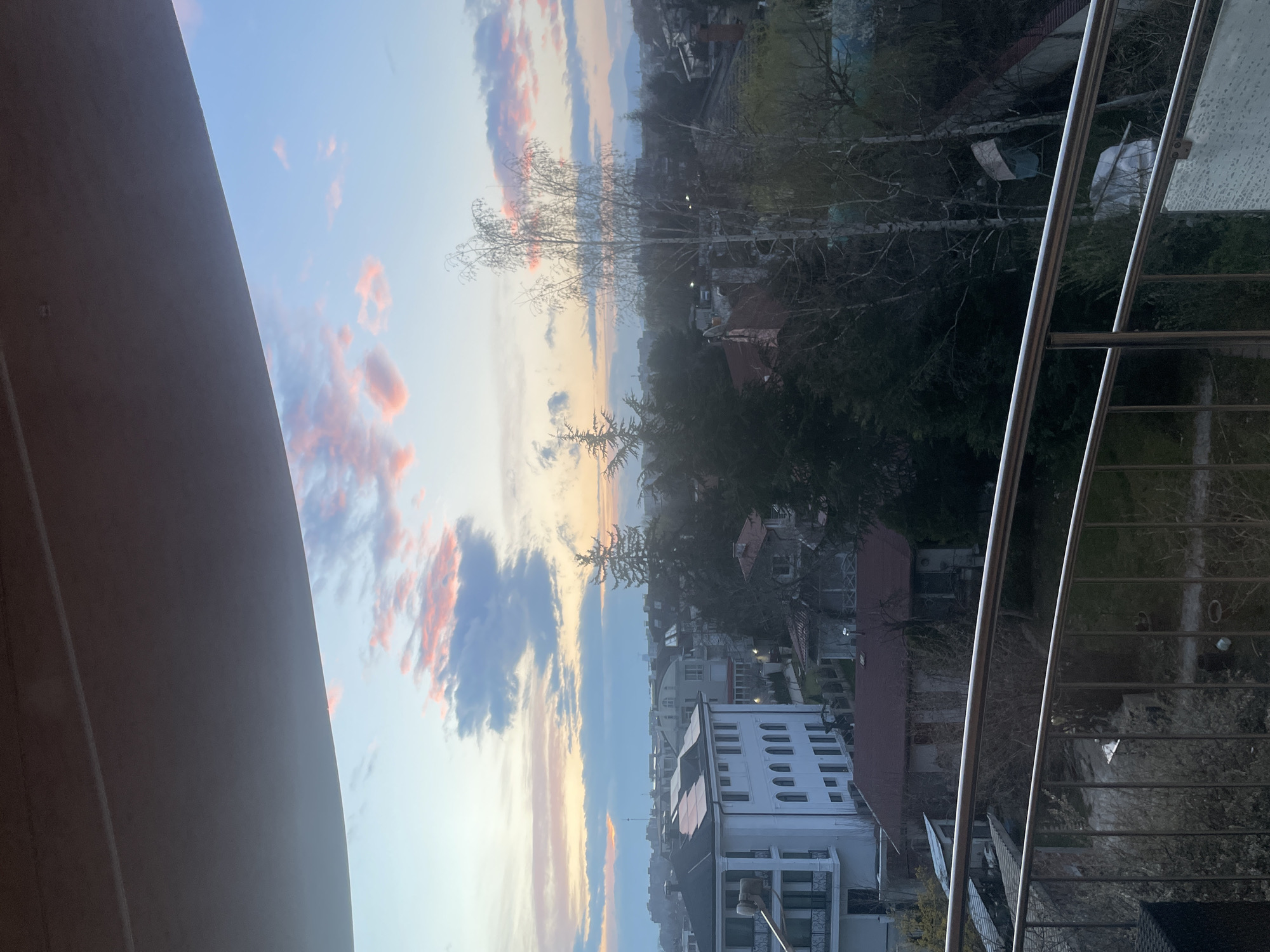 An elevated, static vantage point from a balcony captures a residential landscape at twilight in the city of Unknown, Unknown. The upper portion of the frame is dominated by a sky exhibiting a gradient from light blue to pale orange and pink, with scattered grey and pink-illuminated cumulus clouds. Distant mountains are discernible on the horizon.
The midground reveals a dense urban or suburban environment characterized by numerous buildings and vegetation. A prominent white, multi-story building, equipped with solar panels on its flat roof and featuring multiple windows and balconies, is situated on the left. A white cylindrical object with a metallic arm extends from one of its balconies. Surrounding this structure are various residential houses, many with tiled roofs, interspersed among a mix of bare deciduous trees and dark evergreen conifers. A tall, slender tree with light-colored bark and no leaves stands in the right-center midground.
The immediate foreground displays elements of the balcony structure from which the image was captured. The top edge is defined by the curved, light-grey underside of an overhead architectural element. A polished metal railing, consisting of multiple horizontal bars and vertical supports, spans the lower third of the frame. A dark, woven outdoor furniture piece is partially visible on the bottom left, and a section of translucent glass secured by metal brackets is present on the bottom right. No individuals are depicted, and no specific actions or interactions are observed.