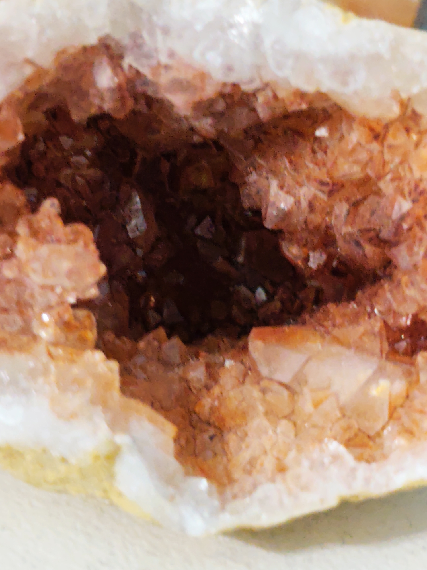 The image presents a detailed close-up of a fractured mineral specimen, identified as a geode. The central feature is an exposed cavity lined with numerous crystalline formations. These crystals vary in size and clarity, exhibiting a predominant color palette of deep orange, amber, and reddish-brown, with some lighter, more translucent facets reflecting light, indicative of quartz or a similar mineral. The crystalline structures are sharply pointed and tightly packed, creating a textured, shimmering surface within the cavity. The interior of the geode appears darker and more recessed, suggesting depth. Surrounding the vibrant crystal core is a visibly contrasting outer matrix, predominantly white to off-white, with segments of pale yellow along the bottom edge, characteristic of the host rock. The overall texture of the specimen is rough and irregular. This mineral specimen is located in the city of Pazardzhik, Bulgaria.