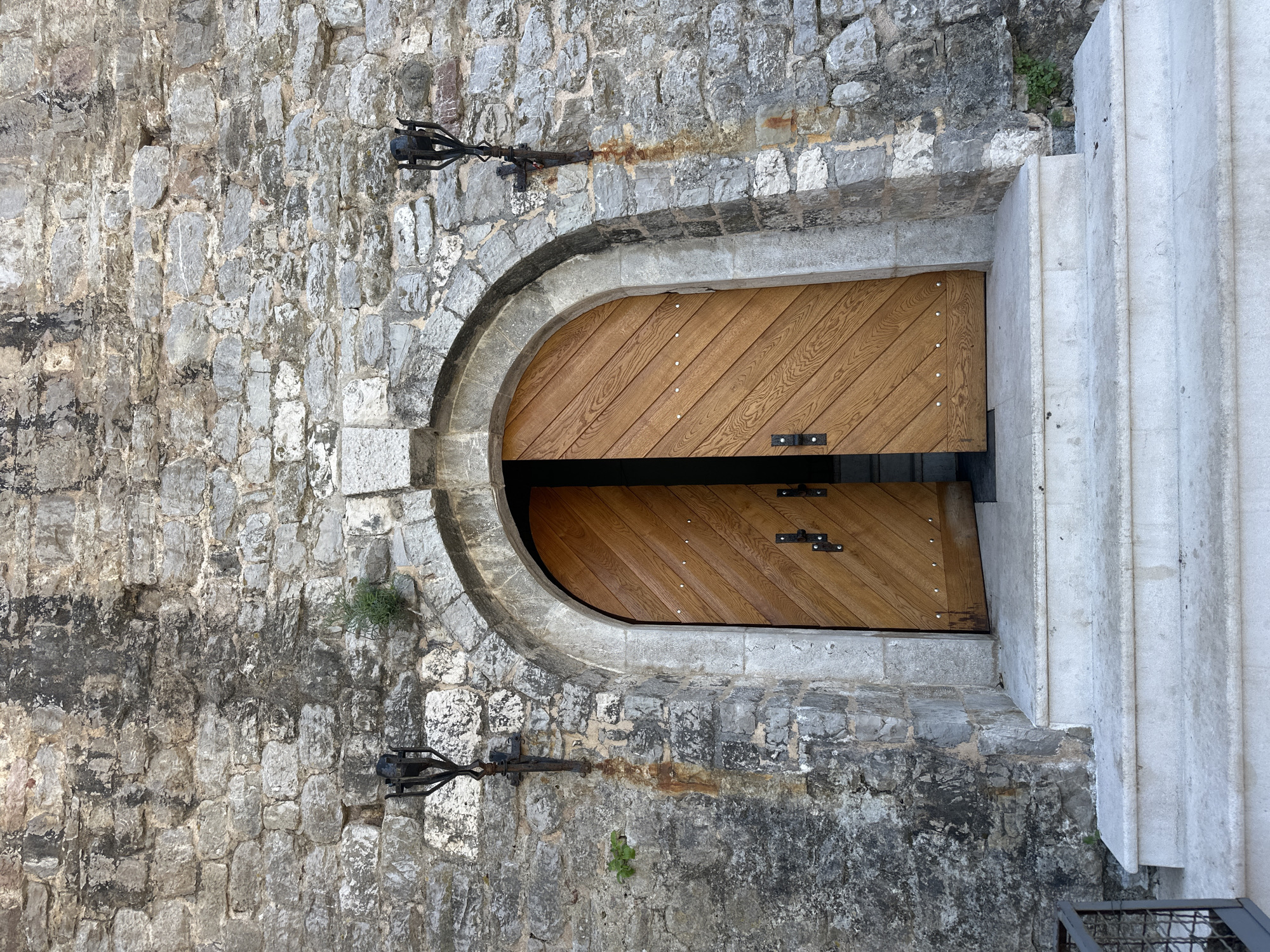 The image depicts a partially opened wooden double door set within a stone archway in the city of Budva, Montenegro. The wall surrounding the archway is constructed of rough-hewn stones, and two rusted metal torch holders are mounted on either side of the arch. The door is made of diagonally patterned planks and has metal hinges and latches. Next to the stone archway is a set of modern white concrete steps.