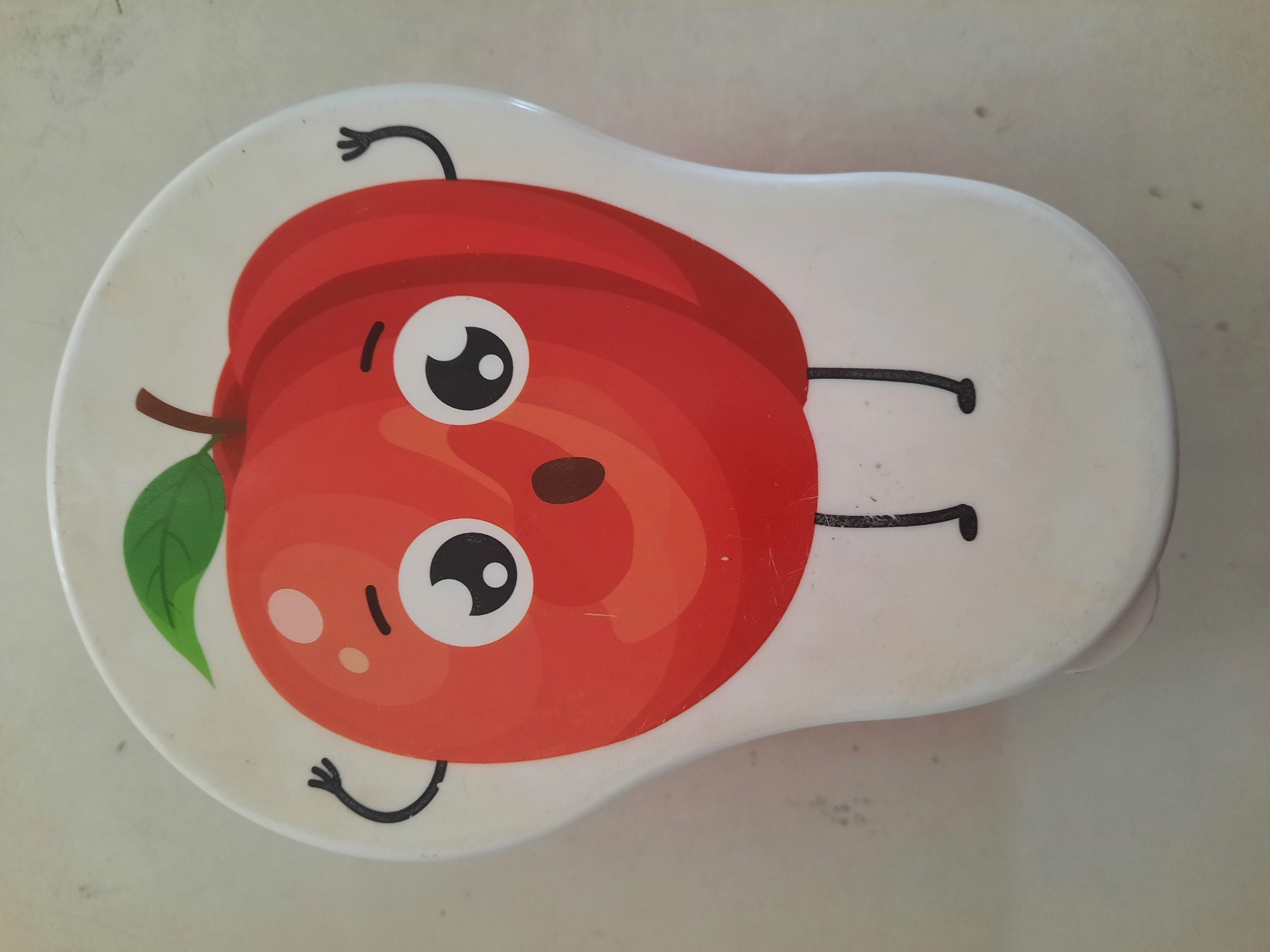 The image shows a cartoon apple character on a white, oval-shaped object. The apple is red with a green leaf and a brown stem. It has large, wide eyes, a small mouth, and two black stick arms raised in a surprised gesture. Two thin black legs extend from the bottom of the apple. The object appears to be a lid or a container, resting on a light-colored, textured surface, likely a table or countertop. There are a few small dark specks on the surface. The lighting is bright and even, casting minimal shadows. There are no people or animals visible. The location is described as being in the city of Lhokseumawe, Indonesia, though there are no visual cues in the image to confirm this.