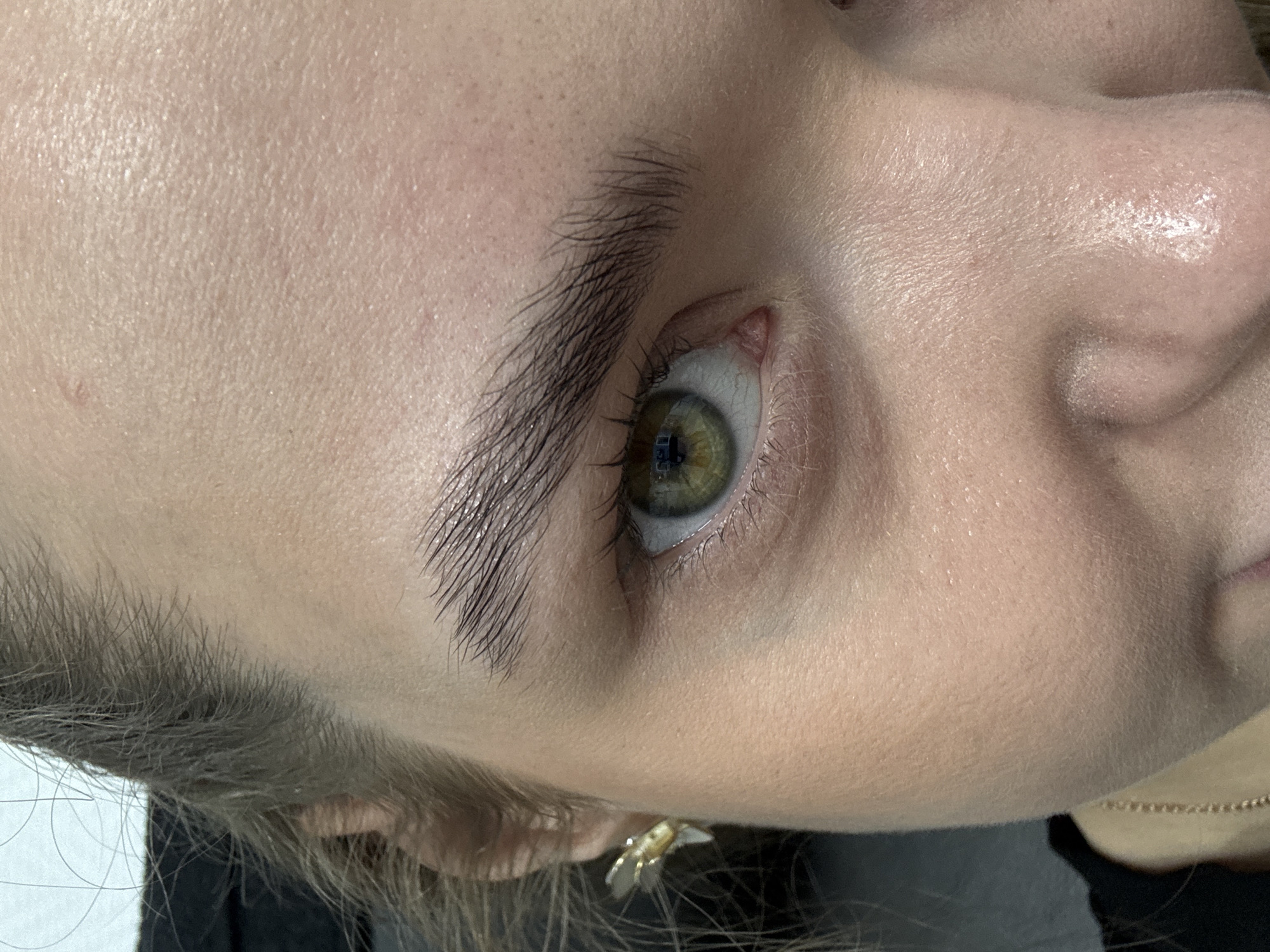 Close-up view of a person's face featuring a green eye with brown and yellow accents, thick dark eyebrows, a nose, and a mouth partially visible. The skin texture is visible, showing pores. An earring is on the ear. A fine gold chain is around the neck. The image is cropped and angled, so the orientation is not traditionally upright. No indications of the location, city, or country are visible in the picture.