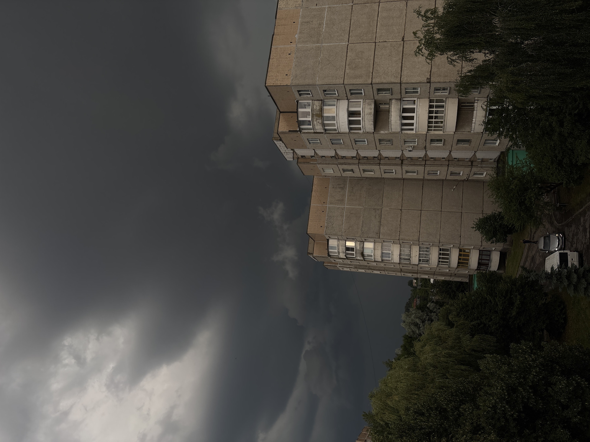 The image depicts a cloudy, overcast sky with dark storm clouds dominating the upper left portion of the frame. Two multi-story residential buildings, constructed with concrete panels, are visible on the right side, extending from the top to the bottom of the image. The buildings have a series of regularly spaced windows and balconies. To the right of the buildings, dense green trees and foliage are present, with a small patch of grass and what appears to be a road or pathway. A white van and a silver car are parked near the buildings. One person, dressed in dark clothing, is standing near the vehicles. The overall mood is somber, likely due to the dark, stormy weather. The scene appears to be set in a residential area, possibly in Lida, Belarus, given the context. The time of day is unclear due to the heavy cloud cover. There is no visible text in the image.