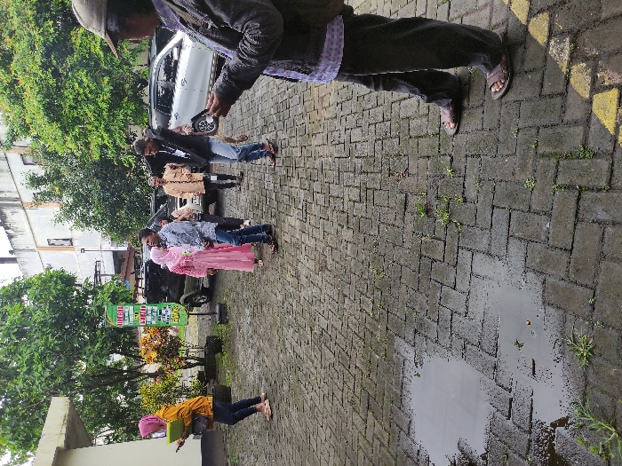 The image depicts an outdoor scene featuring a line of people waiting on a paved area, suggesting a community gathering or service point in Kepanjen, Indonesia. The ground, made of grey interlocking bricks, is visibly wet with several puddles, indicating recent rain. The sky appears overcast, suggesting it is daytime.

In the foreground, a person wearing a hat, dark jacket, and sandals stands, looking down at what appears to be a phone or device. Behind this individual, a queue of several people stretches into the midground. This group includes men and women dressed in casual attire, with some individuals wearing traditional Indonesian garments like a headscarf (hijab) and patterned shirts. One woman in the line is prominently featured in a pink dress and matching headscarf. Further to the left, another person wearing a yellow top and pink headscarf is seen bending over, also looking down at an object in their hands, possibly a book or electronic device.

In the background, a white SUV and a dark truck are parked. Lush green trees and shrubs provide natural scenery, and parts of multi-story buildings are visible beyond the foliage, indicating an urban or semi-urban setting.

A prominent green sign stands on the left side of the image. Clearly visible white text at the top reads "POSKO". Below this, "PEDULI COVID-19" can be partially deciphered, followed by what appears to be "PENERIMAAN BANTUAN" further down. This text suggests the queue is likely for a COVID-19 relief or assistance post, where people might be receiving aid or support. The overall atmosphere conveys a sense of patient waiting for a community service.