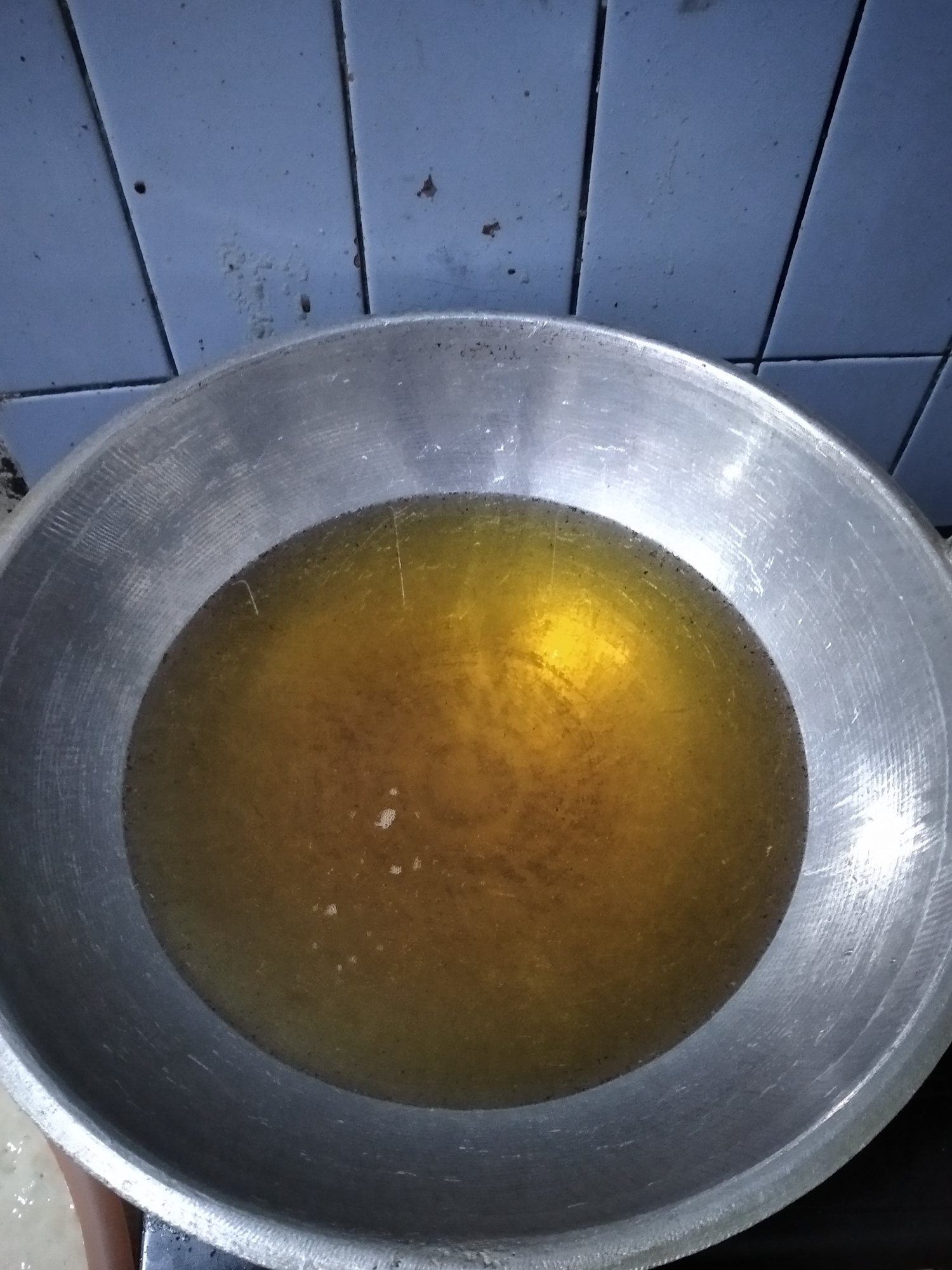 A close-up, overhead shot shows a metal wok filled with hot, golden oil. The wok is resting on a dark surface, possibly a stove. In the background, a wall tiled with light blue squares is visible. The tiles appear somewhat aged and have faint marks and grout lines. The oil in the wok has a slight sheen and a few small bubbles, indicating it is hot. The overall scene suggests preparation for frying food. The specific location in Rambipuji, Indonesia, is not discernible from the visual elements.