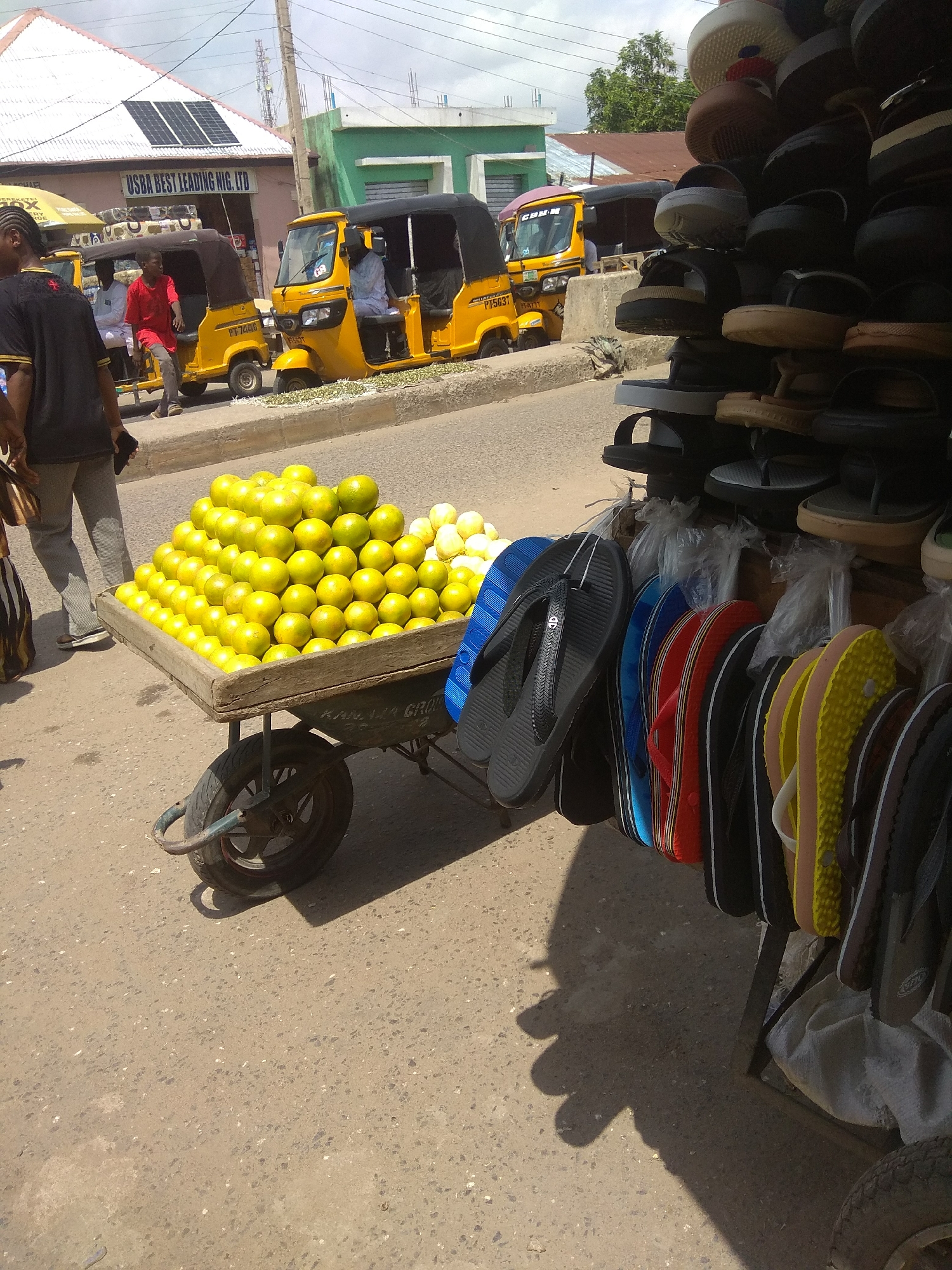 The image depicts a street scene in Jalingo, Nigeria, likely a marketplace. In the foreground, a wooden cart is laden with a large pile of green apples. To the right of the cart, various colorful flip-flops and sandals are displayed on a rack, some still in plastic packaging. Behind the cart, two yellow auto-rickshaws are parked on the asphalt road, one occupied by a person in white clothing and the other with a driver in a red shirt and grey trousers. The backdrop includes buildings with corrugated iron roofs, one with solar panels, and a green painted structure. The sky is overcast. The overall impression is of a bustling street vendor environment.