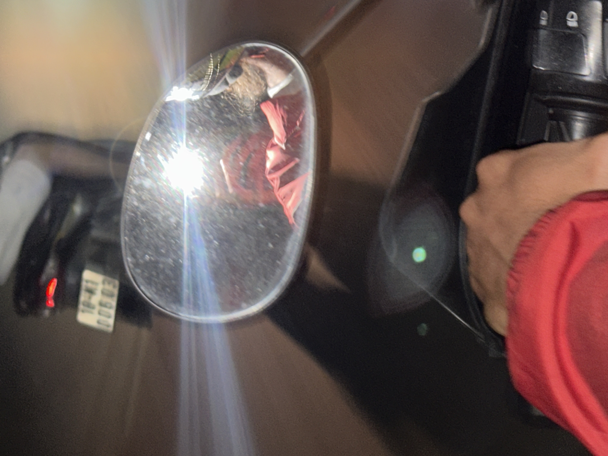 The image is a first-person perspective shot from a motorcycle or scooter in Mai Chau, Vietnam. The rider's hand, wearing a red jacket, is visible in the foreground on the right, gripping a handlebar. A rear view mirror is prominently displayed in the center, reflecting the rider, who is wearing a red jacket, sunglasses, and a helmet with gold trim. The license plate of another vehicle, which reads "18-K1 006 03", is visible on the back of a scooter in the background to the left, along with its red taillight. The scene appears to be on a street at night, indicated by the reflections and light flares, typical of urban or semi-urban environments in Mai Chau.