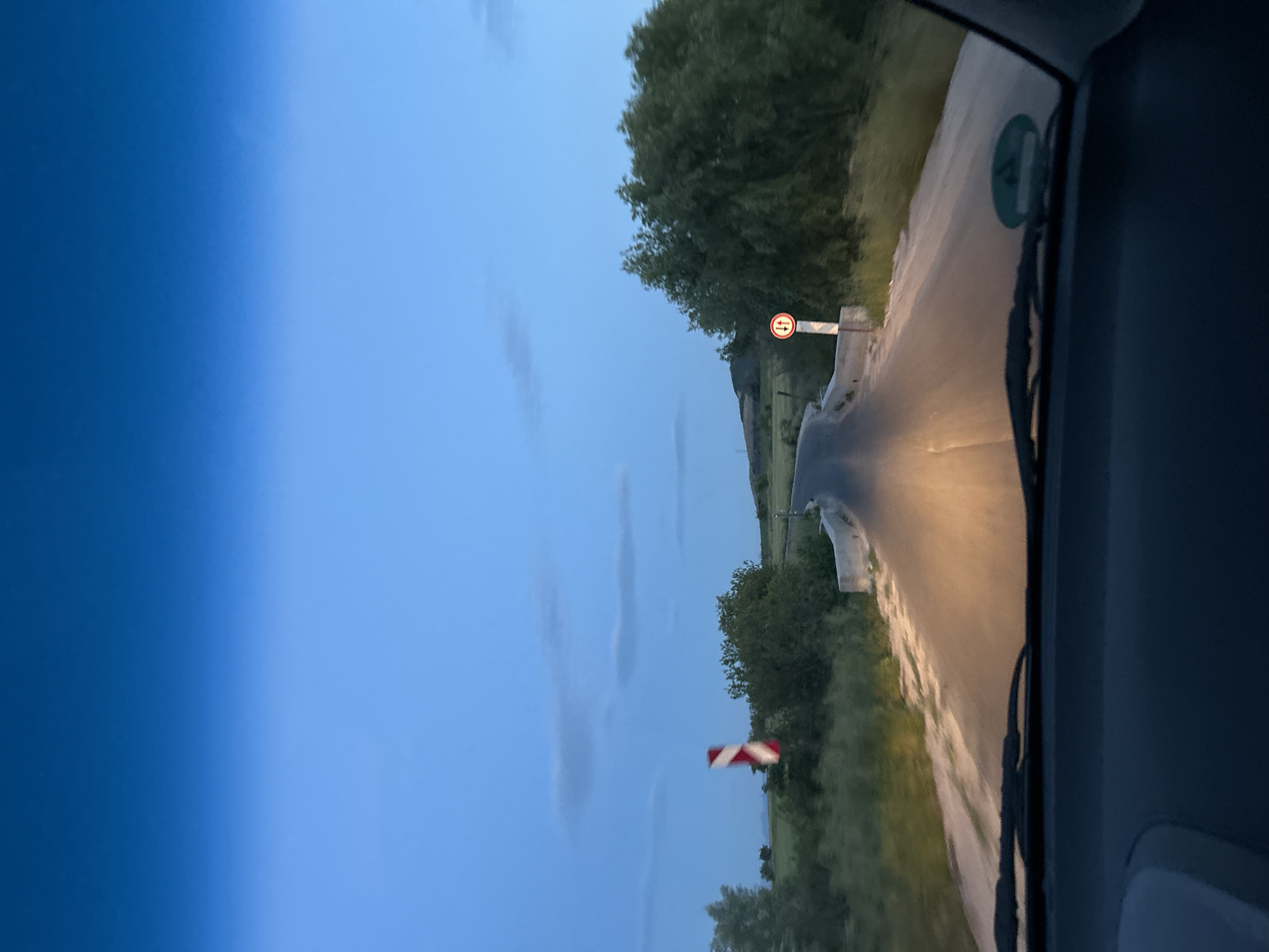 The image shows a view from inside a vehicle driving on a rural road. The sky is overcast, suggesting either dawn or dusk. On the right side of the road is a bridge with a traffic sign indicating oncoming traffic has the right-of-way. Further along the road, there is a railroad crossing signal. The road appears wet. The location might be Ovchepoltsi, Bulgaria.