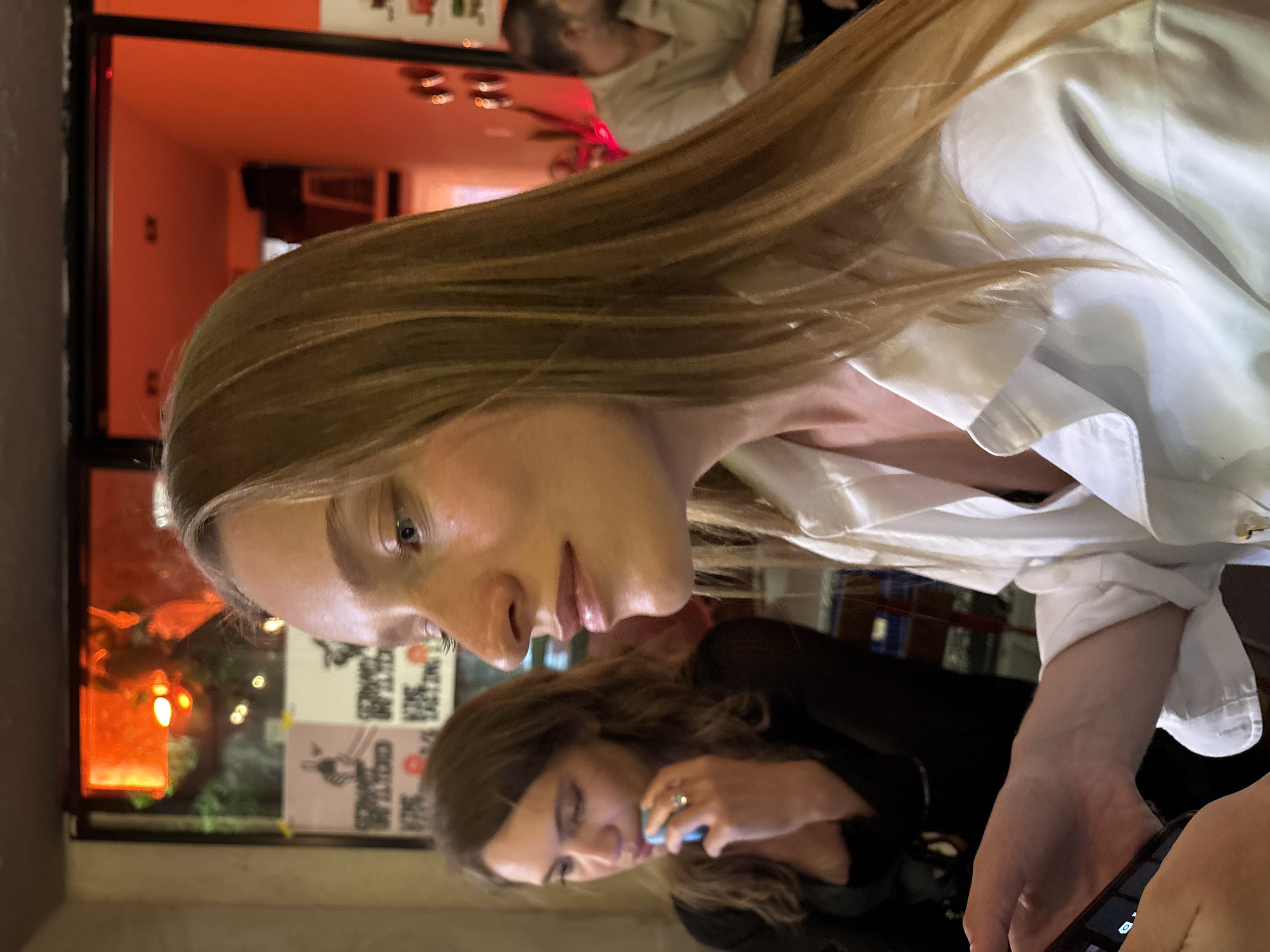 Two women are seated at a table in a dimly lit establishment, possibly a bar or restaurant. The woman in the foreground, facing to the right, has long blonde hair and is wearing a white button-down shirt. She is holding a smartphone in her left hand, her thumb resting on the screen. To her left and slightly behind her, another woman with brown hair is visible. She is wearing a dark top and appears to be holding a small blue object near her mouth, possibly a drink or a piece of food. The background shows posters on the wall and a glowing orange light from an interior space or window. A man in a light-colored shirt is visible in the far background. The overall ambiance suggests a social gathering. This scene is likely from Tel-Aviv, Israel, given the context.