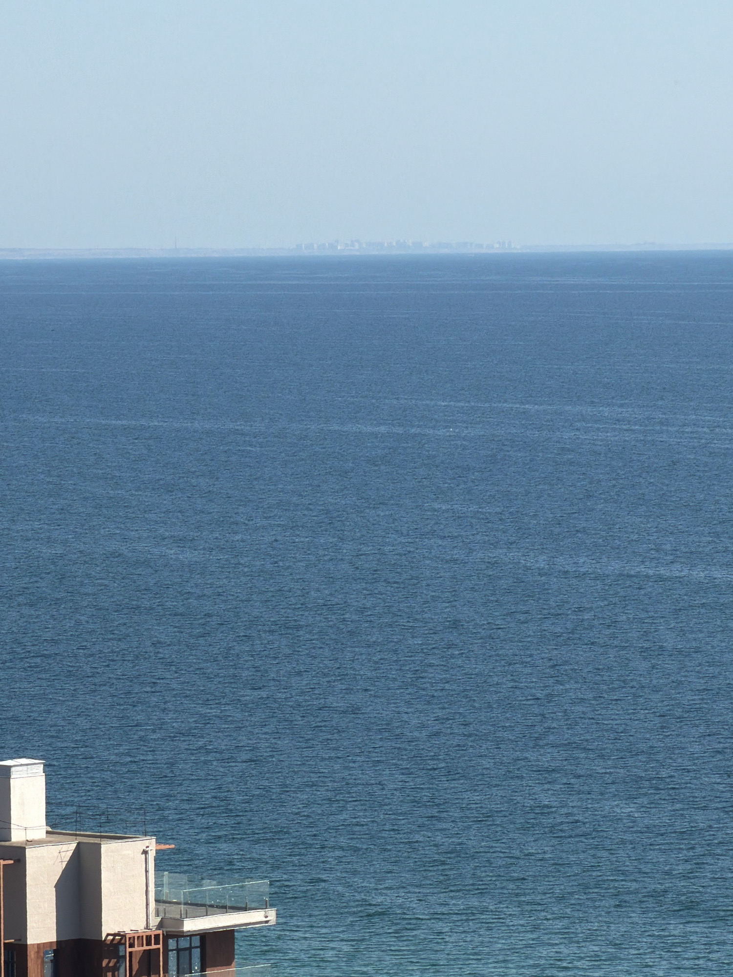 The image shows a wide expanse of blue ocean with a hazy skyline in the distance. In the foreground, the corner of a modern building with a balcony overlooking the sea is visible. The sky is a pale blue and clear, suggesting good weather. The water is calm with gentle ripples. The distant skyline is indistinct due to atmospheric haze, but appears to be a coastal city with buildings. The building in the foreground is light-colored with dark trim and features a glass railing on the balcony. There are no people visible in the image. The scene captures a serene view of the Black Sea coast, likely in Odesa, Ukraine, given the presence of coastal architecture and the characteristic haze often seen over the water.