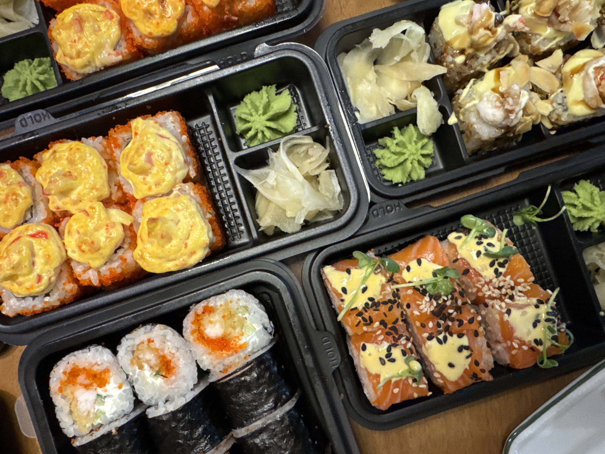 The image shows four black plastic containers of sushi. The containers are arranged on a light brown surface. The sushi appears to be of various types and fillings, including rolls topped with a yellow sauce, rolls with salmon, rolls with tobiko, and rolls wrapped in nori. Each container also includes portions of wasabi and pickled ginger. There is no indication of Tomilino, Russia or any people, actions, or interactions in the image.
