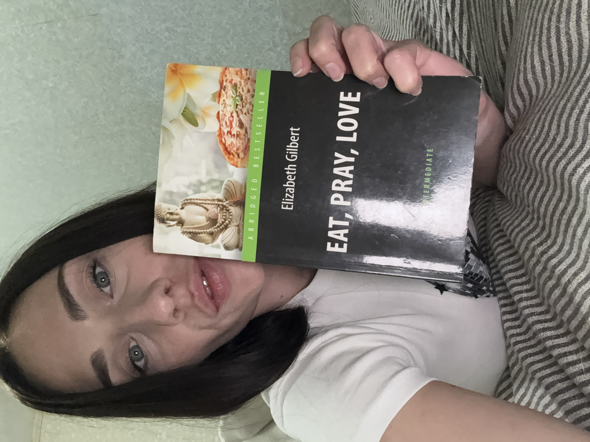 A woman is holding a copy of Elizabeth Gilbert's "Eat, Pray, Love" book in front of her face. The book cover features an image of a Buddha, flowers, and pizza. The woman has dark hair and light-colored eyes. She is wearing a white t-shirt. The background includes a light-colored wall and a striped pillow or blanket. There is no information to determine the location is Saint Petersburg, Russia.
